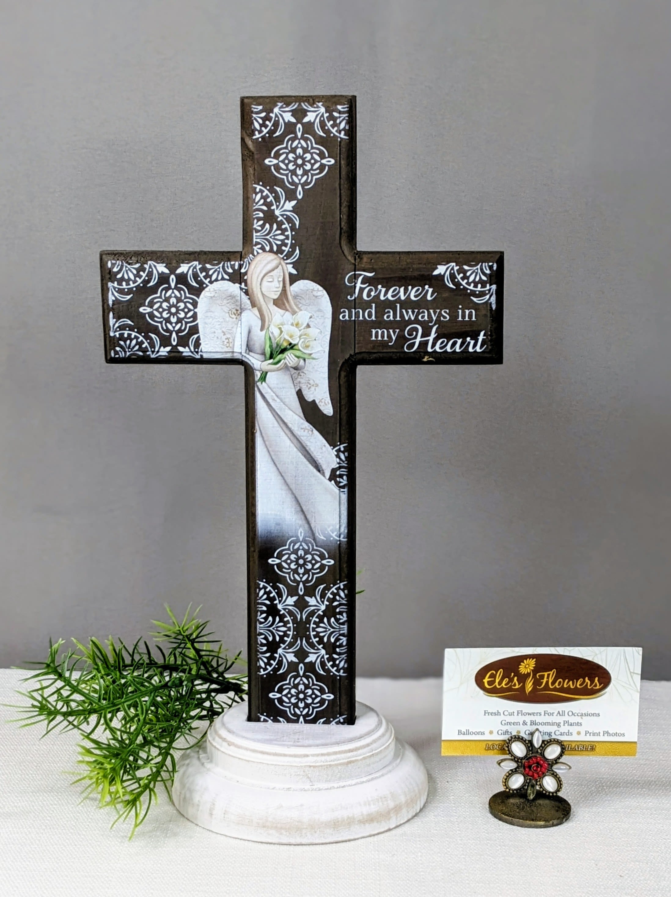 Wooden Angel Sympathy Cross - This beautifully crafted wooden cross features a gentle angel holding white lilies, symbolizing peace, love, and remembrance. Set against a rich wood-toned background with elegant white detailing, it is accented with the heartfelt message: “Forever and always in my heart.”  Designed to offer comfort during times of loss, this cross rests on a sturdy, softly distressed base, making it perfect for display on a table, shelf, or memorial space. Its timeless design and soothing imagery create a meaningful tribute to honor a cherished loved one.  A thoughtful sympathy gift that expresses love, faith, and lasting remembrance when words are not enough.  Approximate Dimensions: 13"H x 8"W
