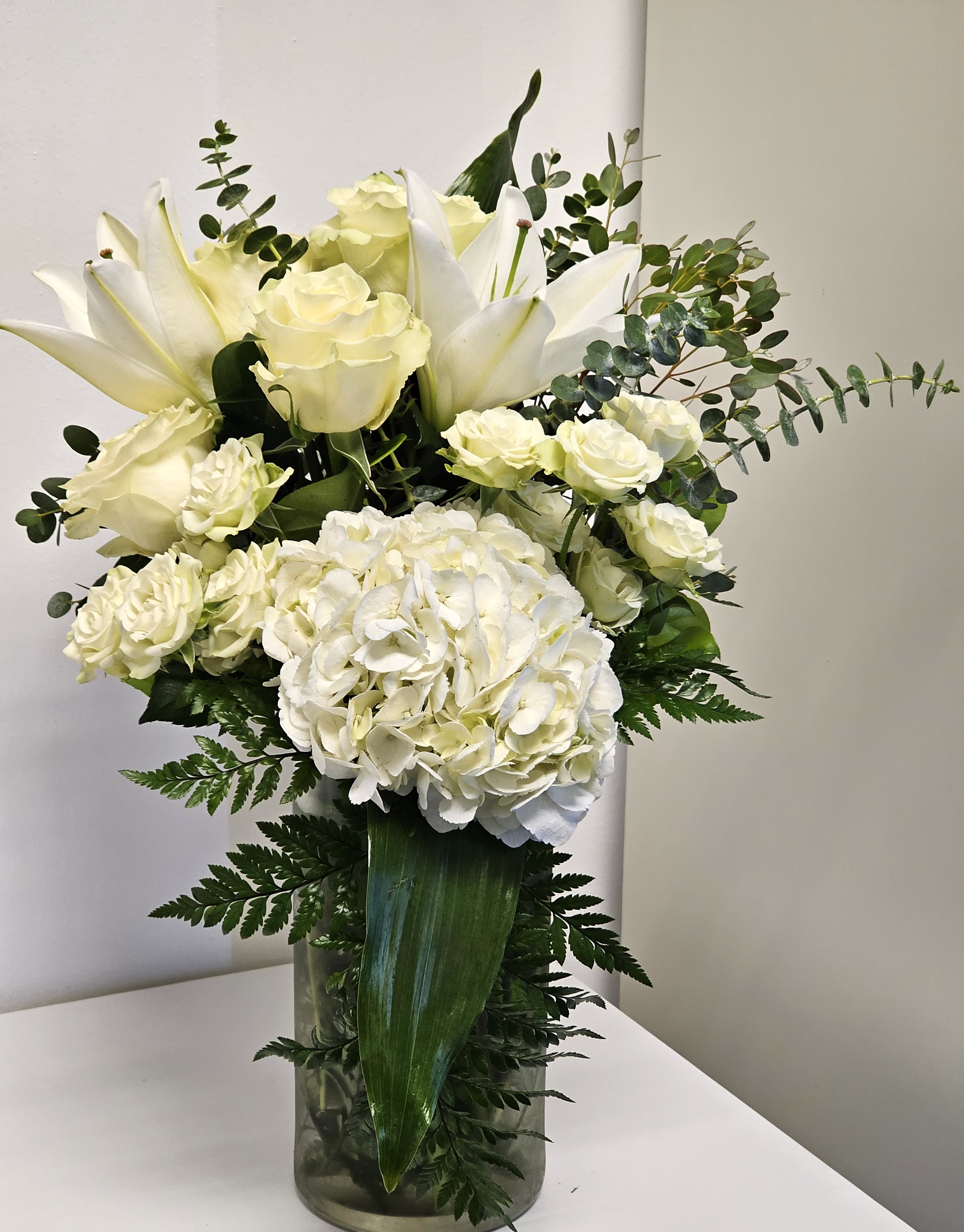 White arrangement with white roses -   This exquisite all-white arrangement embodies grace and sophistication. At the heart of this design are pristine white roses, symbolizing purity and love. Surrounding the roses are complementary white blooms such as delicate lilies, fragrant stock, and fluffy hydrangeas, creating layers of texture and depth. Soft, silvery eucalyptus leaves and other light greenery are subtly woven throughout to enhance the natural beauty of the flowers without overpowering their elegance. Presented in a sleek, white ceramic vase, this arrangement is a timeless choice, perfect for weddings, anniversaries, or any occasion where elegance is key.  