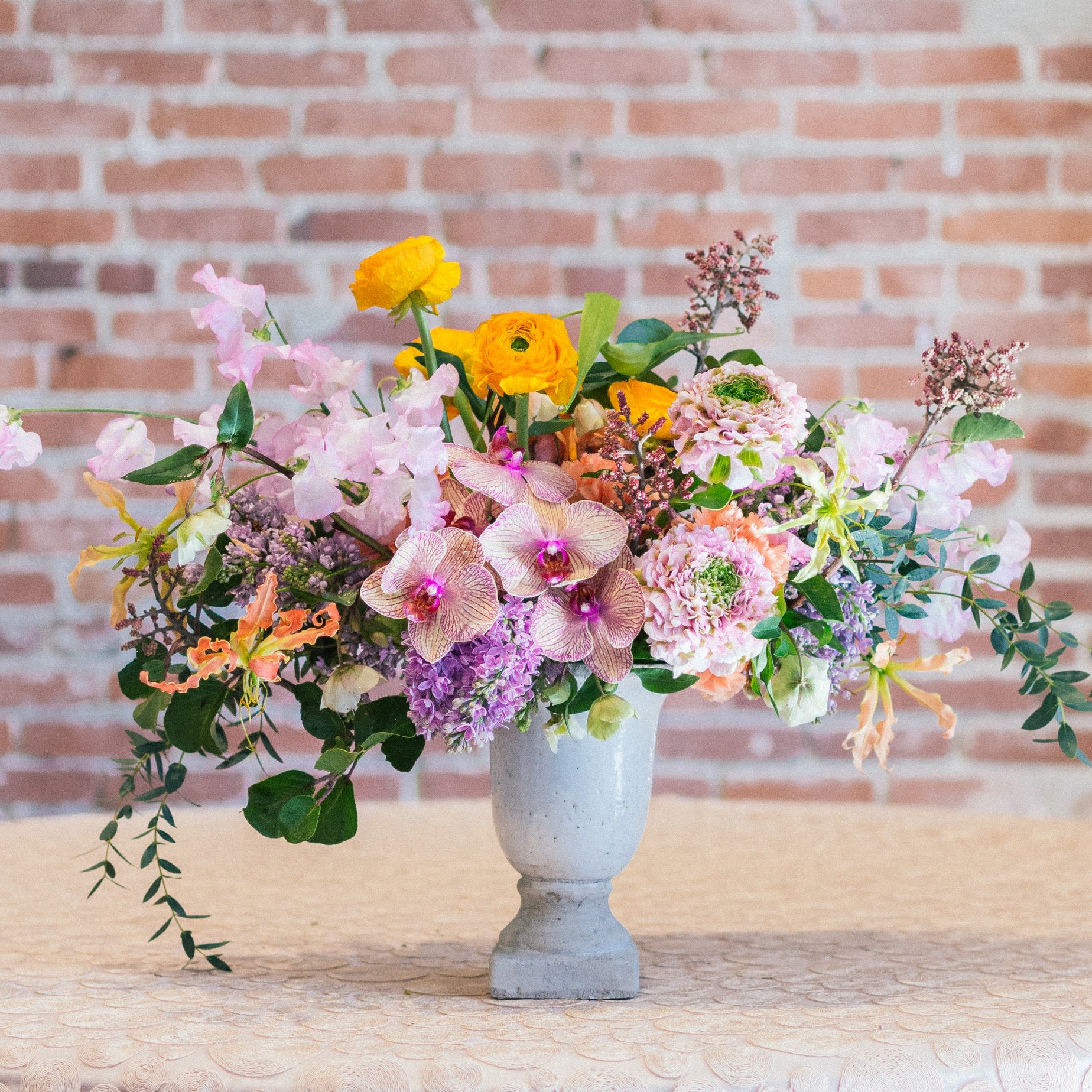 Sweet Love garden bouqy - A lush, garden-style centerpiece featuring golden ranunculus, blush roses, orchids, sweet peas, and cascading greenery. Designed in a modern stone pedestal vase, it’s a romantic statement piece perfect for luxury gifting or elegant tables.