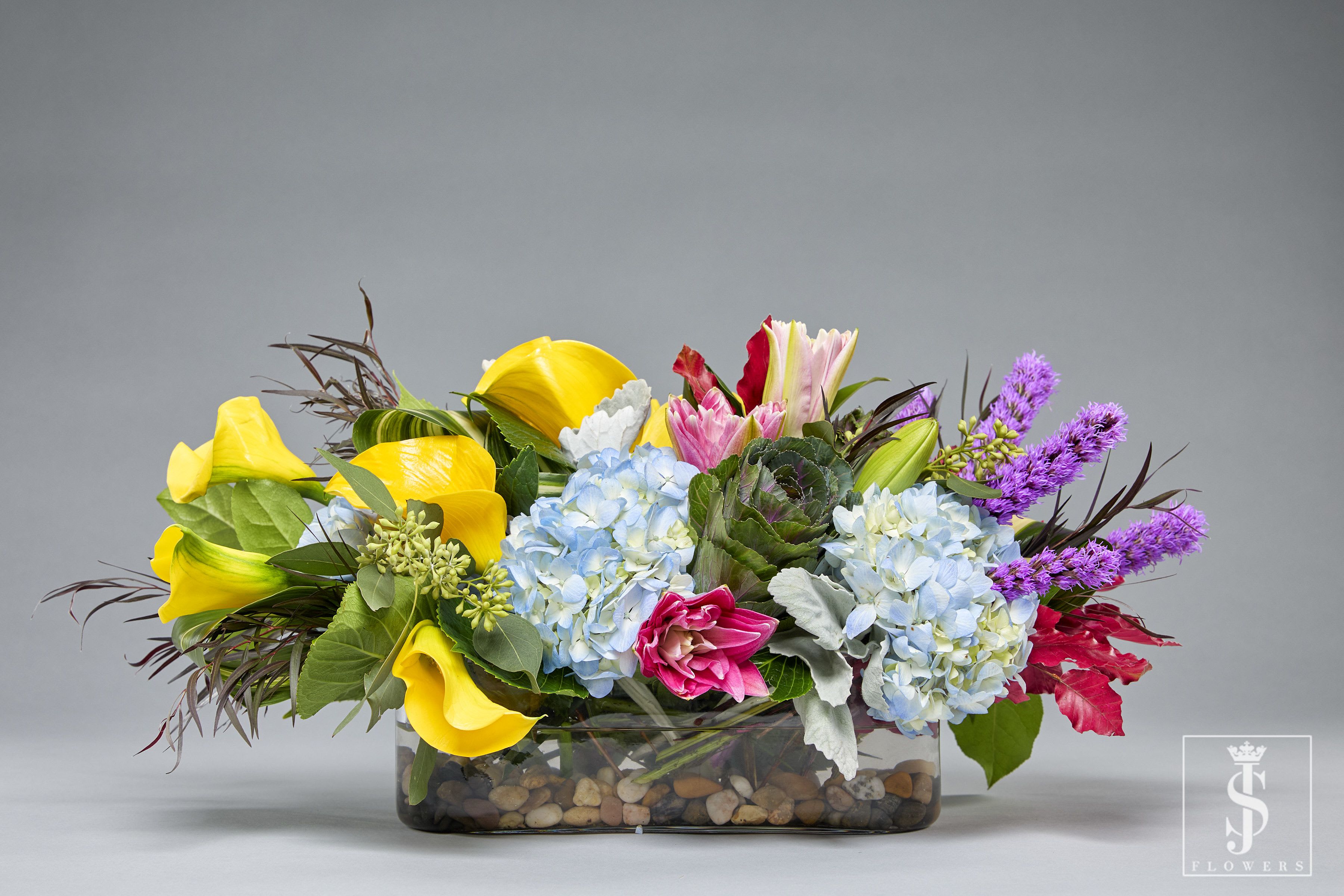 Pastel Elegance Centerpiece - This vibrant centerpiece captures the essence of spring and summer with a brilliant mix of colors and textures. The cheerful yellow calla lilies radiate warmth and joy, while the serene blue hydrangeas add a sense of tranquility. Soft pink tulips and lilies bring a touch of romance and charm, perfectly complemented by the striking purple liatris spikes for a bold and modern accent.  Rich greenery and ornamental kale add depth and structure to the arrangement, with leaves that create a natural, garden-inspired aesthetic. The arrangement is designed in a glass vase lined with decorative pebbles, grounding the design in earthy elegance. Perfect for weddings, celebrations, or as a standout home centerpiece, this creation exudes vibrancy and sophistication.