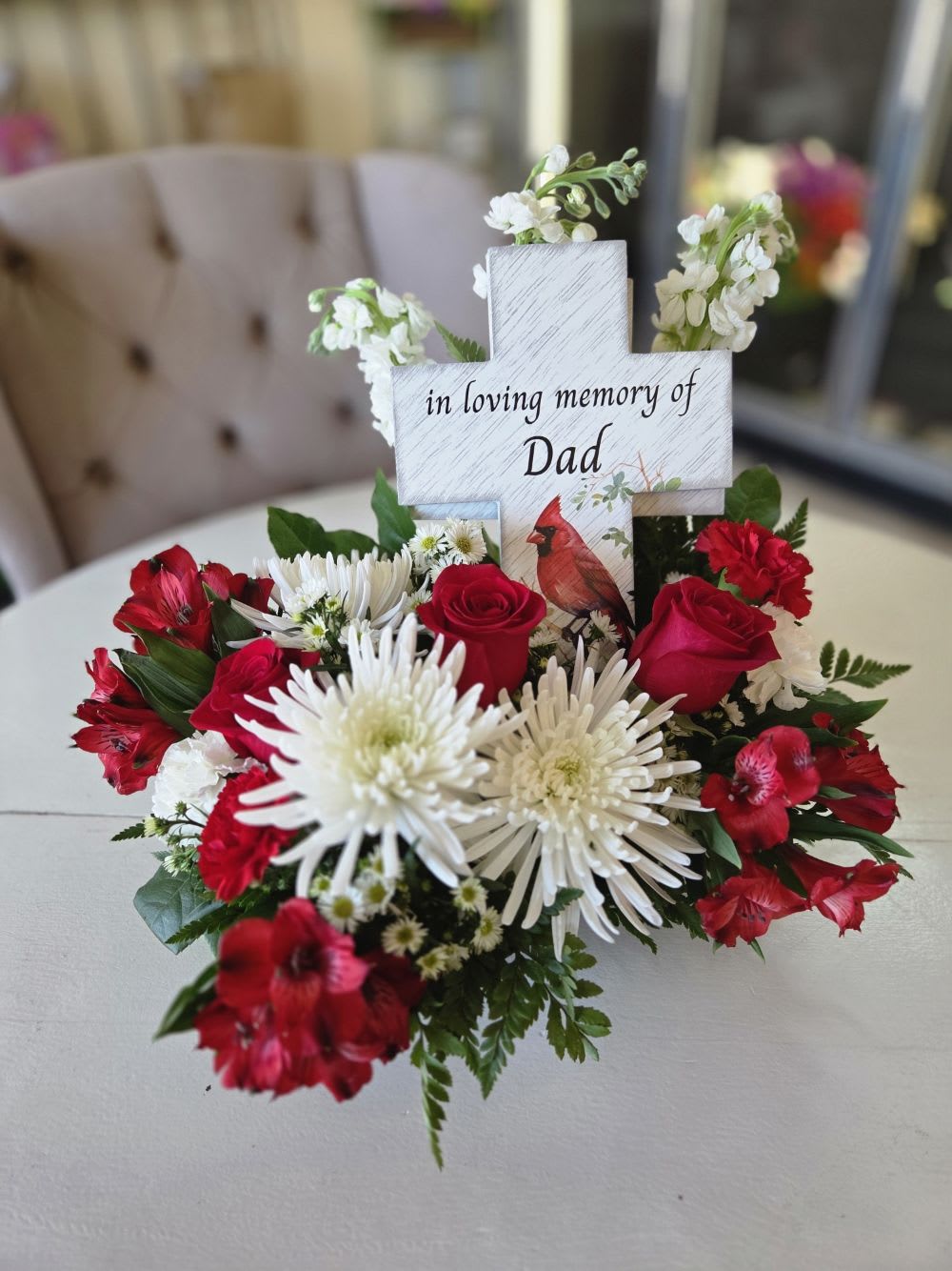 Honor Thy Father - Honor a cherished father’s memory with this elegant and heartfelt floral arrangement. Featuring a beautiful mix of red roses, white spider mums, carnations, and alstroemeria, this tribute combines rich color and graceful texture to express enduring love and remembrance. At the center stands a wooden cross accent inscribed with “In loving memory of Dad” and a red cardinal illustration — a timeless symbol of hope and presence.  Perfect for memorial services, graveside tributes, or home remembrance displays, this arrangement offers a comforting reminder that love lives on.  Approximate Size: 14" H x 16" W Flower Types: Red Roses, White Spider Mums, Alstroemeria, Carnations, Seasonal Greens