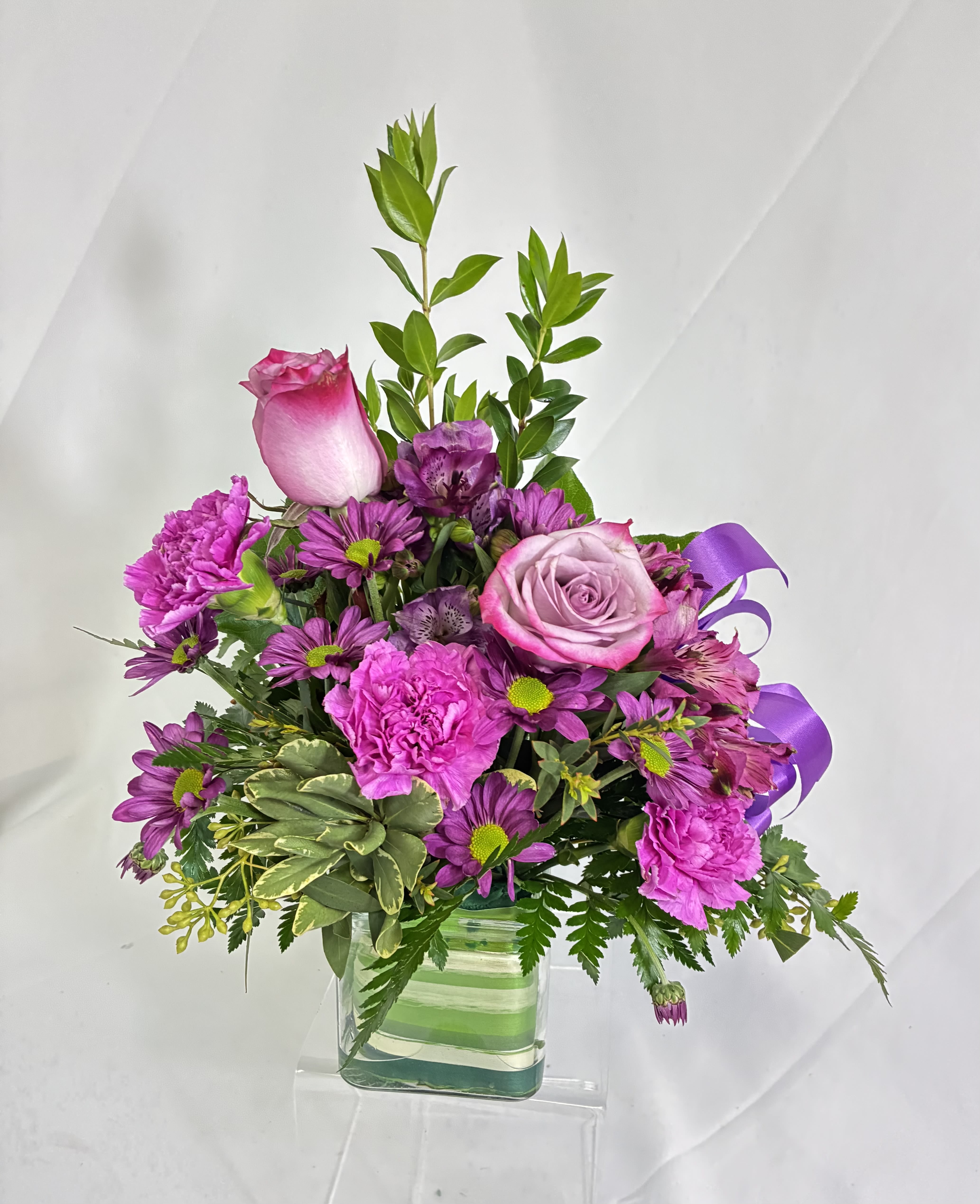 Fancy Floret Cube - A purple lover's dream of Roses, raspberry Daisies, lavender Alstroemeria, Chrysanthemums and accented with purple Monte Casino blooms, and greenery.  This fresh purple floret is presented in a glass cube, accented with Aspidistra leaf, for a display of this purple &amp; lavender arrangement.