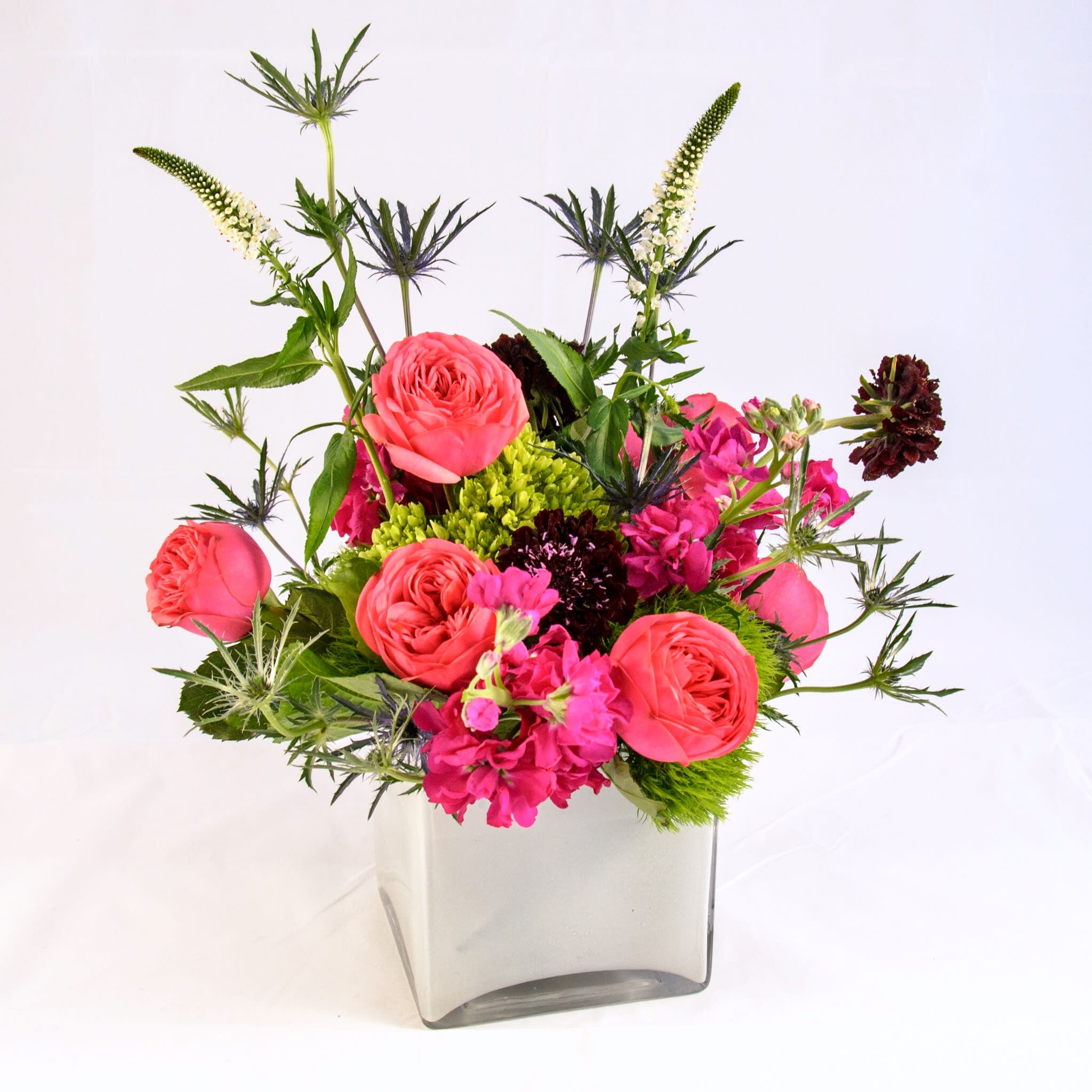 Isabelle - Jewel-tone, enriched color with fuchsia pink roses, fragrant stock, mini green hydrangea, blue thistle, white veronica, and a touch of deep purple scabiosa. Low-compact arrangement in modern white glass cube vase. Shine bright in any occasions. (Approx. size 10"w x 12"h x 10"d)