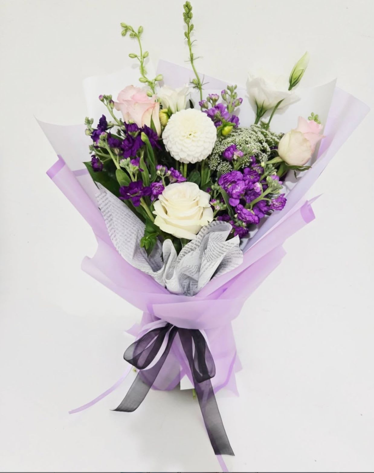 Lavender Haze™ - Lavender Haze evokes that fleeting moment between dreaming and waking—where feelings are soft, intentions are pure, and everything is tinted in lilac. Featuring creamy white dahlias, blushing pink roses, and clusters of violet blooms, this bouquet is a study in restrained romance. Wrapped in sheer lilac and black ribbon, it's designed for those who love with quiet intensity.
