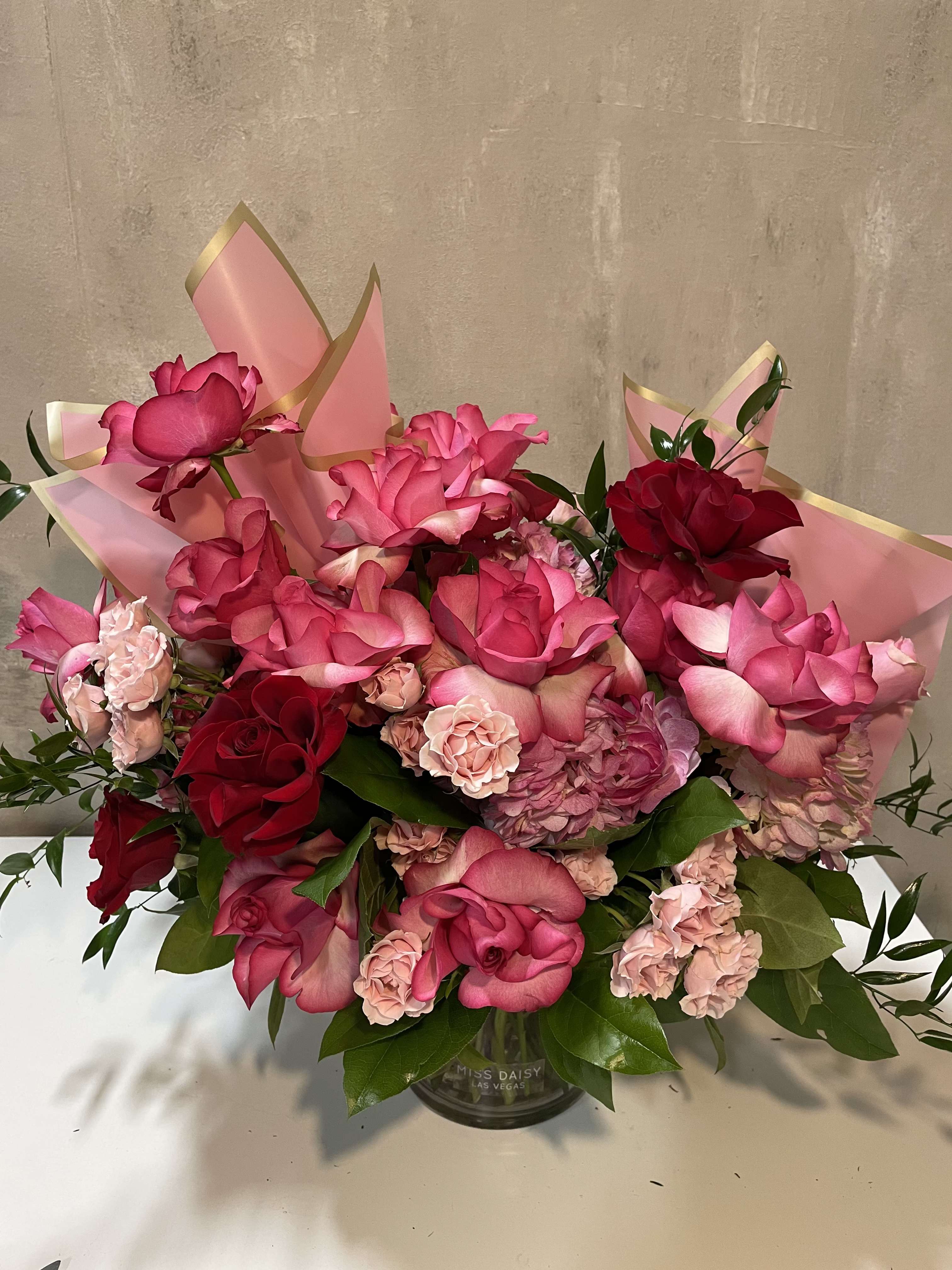Blushing Romance  - This hand crafted premium floral arrangement is  lush and feminine featuring premium garden roses in shades of raspberry, blush, and classic red, accented with delicate spray roses, hydrangea, and fresh greenery. Designed in a clear glass vase and finished with signature Miss Daisy wrapping for an elevated presentation, this piece is perfect for celebrations, heartfelt gestures, or simply making a beautiful statement.