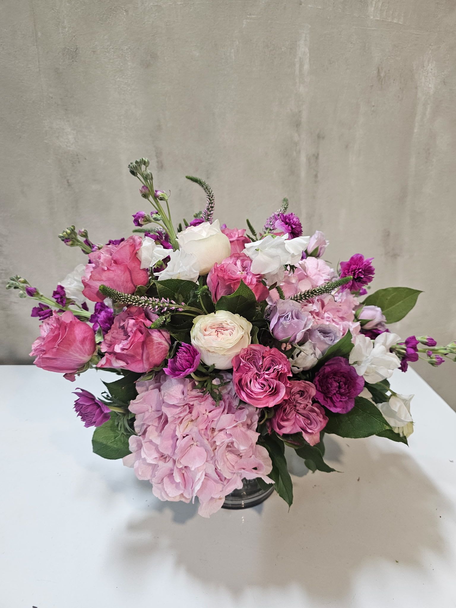 Petal Poise - A graceful garden-style arrangement featuring romantic pink roses, soft hydrangea, delicate seasonal blooms, and fresh greenery, designed low and lush for a naturally elegant look. Sweet, refined, and timeless, this piece brings effortless beauty to any setting.
