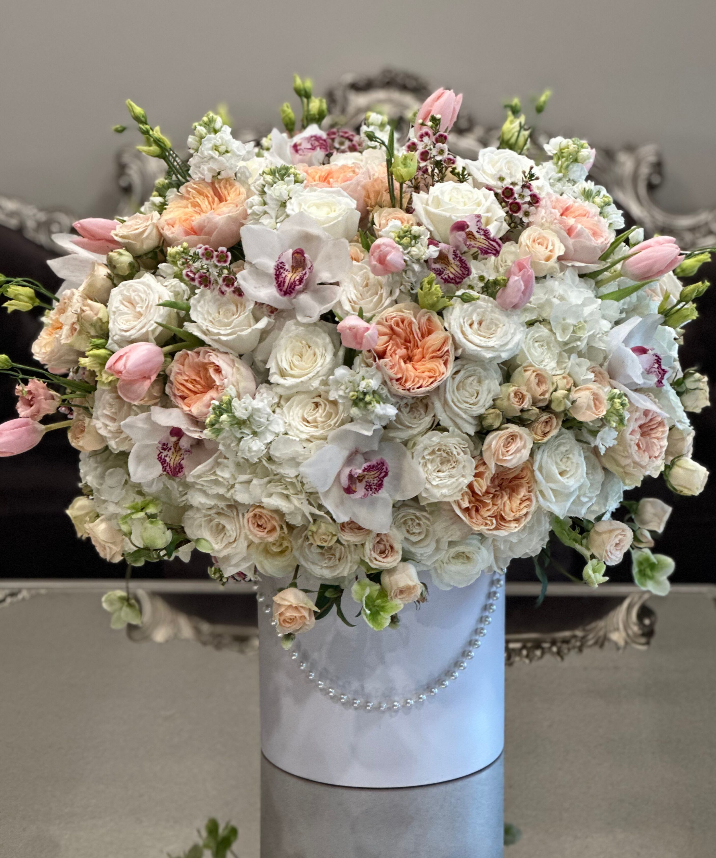The Pearl Box - A luxurious and timeless floral display featuring a pristine blend of white orchids, velvety roses, and delicate tulips. Nestled in a sleek white hat box, each bloom is artfully arranged and accented with soft touches of greenery.