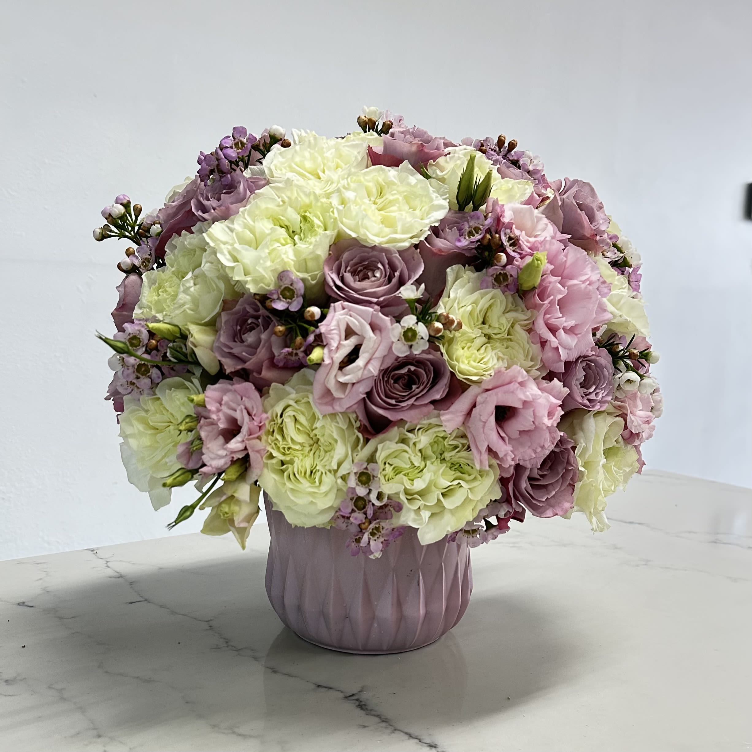 Lavender Haven - A delicate arrangement of lavender roses elegantly placed in a matching lavender vase. The roses, in soft hues of purple, exude a sense of grace and tranquility, with their velvety petals gently unfolding.