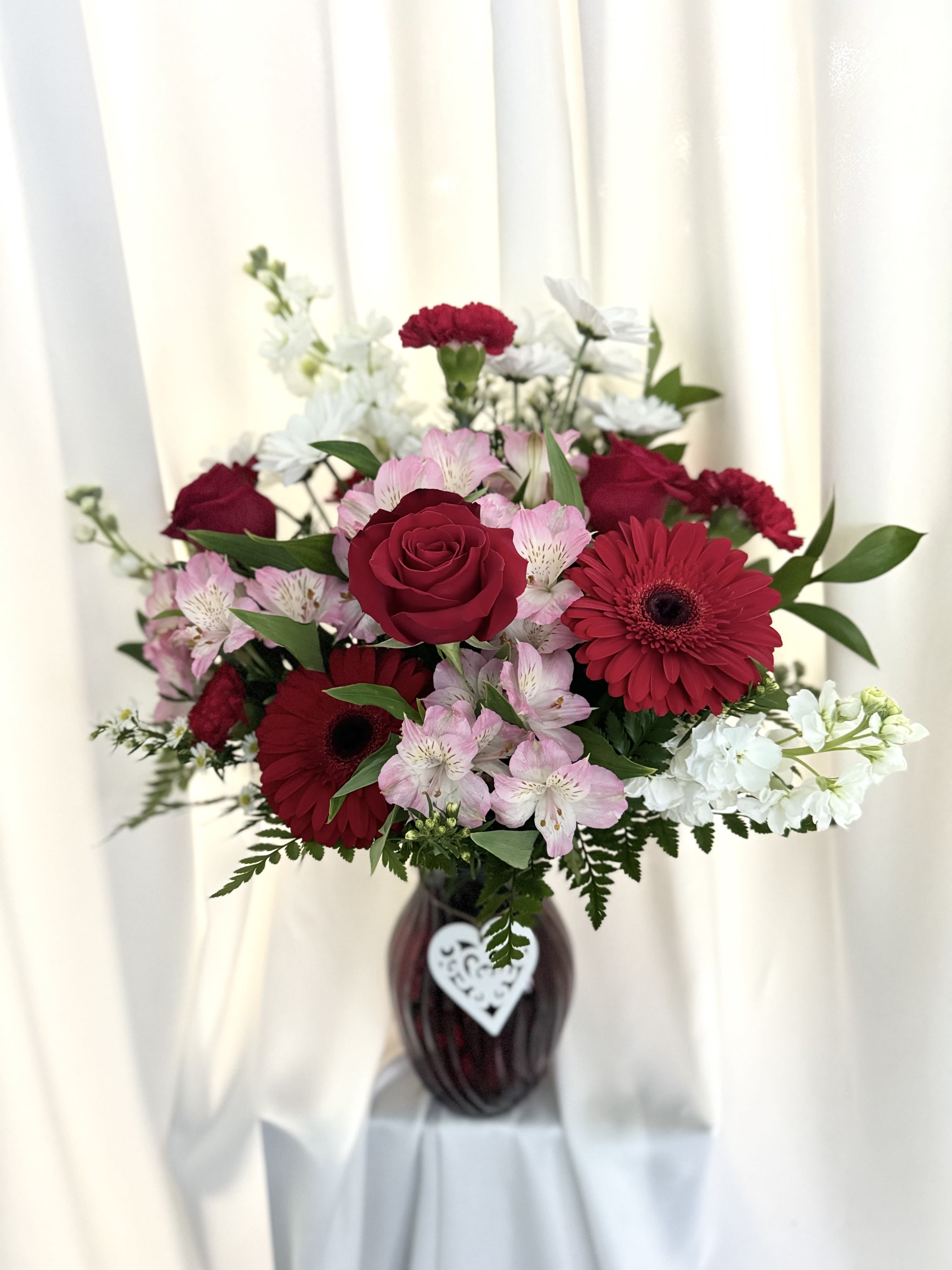 Crimson Romance Bouquet - This stunning arrangement combines rich red roses, vibrant gerbera daisies, soft pink alstroemeria, and delicate white blooms in a deep ruby vase adorned with a heart charm. A perfect expression of love and admiration, this bouquet brings timeless beauty to any celebration or romantic gesture.