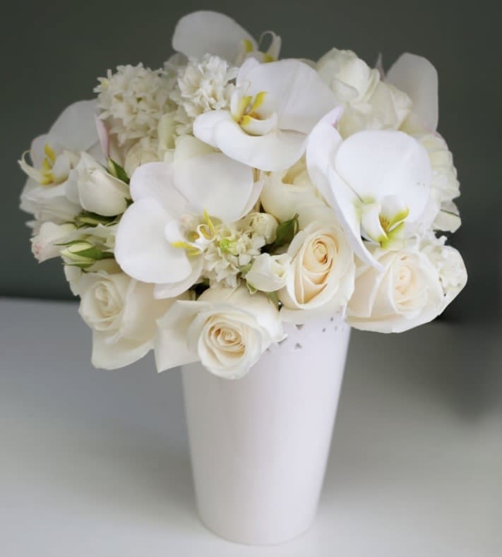 Delphine - This sophisticated arrangement showcases the timeless beauty of white flowers in perfect harmony. Graceful white phalaenopsis orchid blooms take center stage with their smooth, sculptural petals and gentle yellow centers, while cream roses add classic romance throughout. Textural white hyacinth provide fullness and create a cloud-like base that ties everything together.  The monochromatic white palette creates a sense of calm and sophistication that works beautifully in any setting. Each flower type contributes its own character - the exotic elegance of orchids, the timeless appeal of roses, and the soft, textural quality of the supporting blooms. The subtle cream tones add warmth and prevent the arrangement from feeling stark.  Presented in a clean white ceramic vase, this arrangement embodies understated luxury. The simple vessel allows the natural beauty of the flowers to shine while maintaining the pure, sophisticated aesthetic throughout.  Dimensions: - Vase: 4" diameter × 7" height - Overall arrangement: 10-12" height × 8" width  Perfect for weddings, sympathy arrangements, modern spaces, or anyone who appreciates clean, sophisticated floral design in pure white.