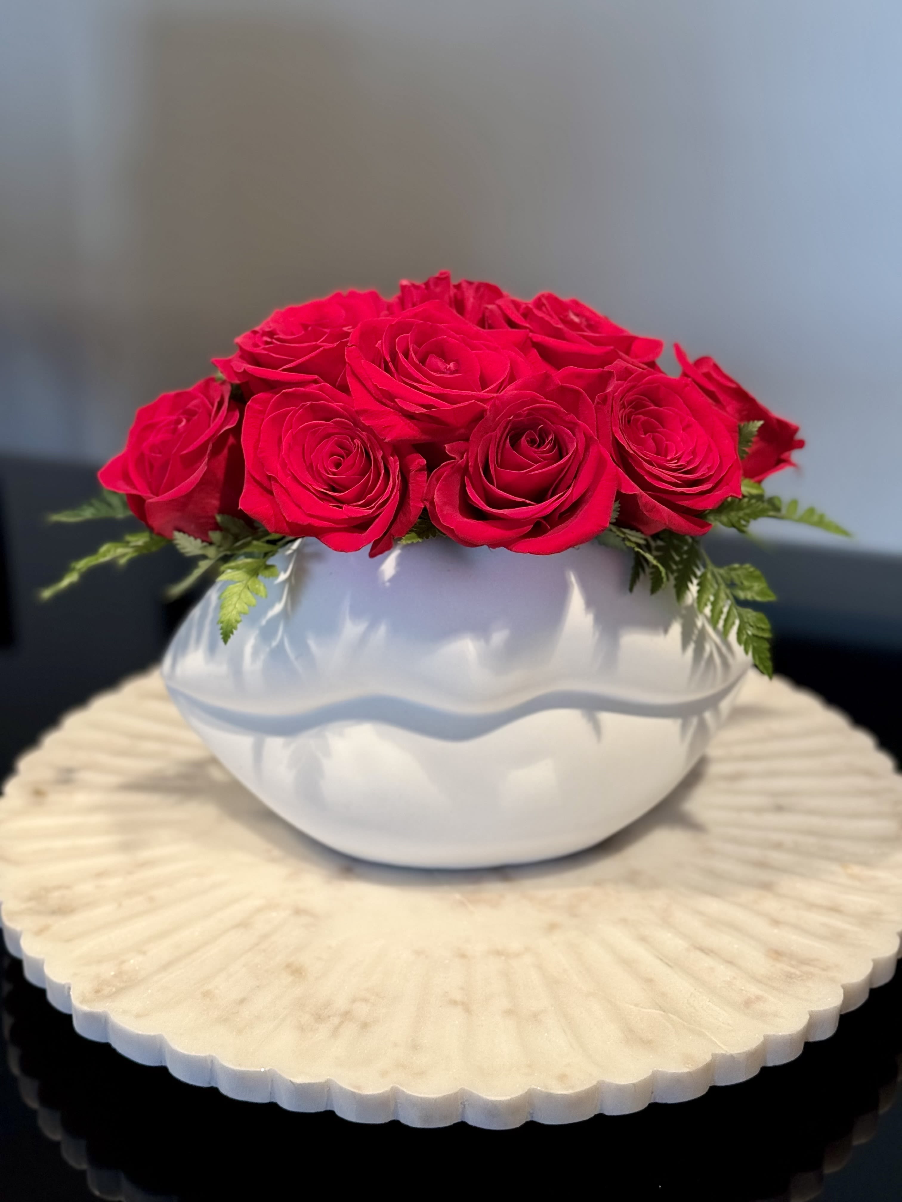 The Muse - A floral arrangement designed to capture hearts. This piece features a lush dome of premium, velvet-textured red roses, accented with delicate sprigs of leather leaf fern for a touch of natural green. The true showstopper is the sculptural matte white ceramic vessel, shaped into a minimalist, stylized pair of lips. Resting on a fluted marble-style base, this arrangement isn't just a bouquet—it’s a conversation piece.  Perfect for: Anniversaries, Valentine’s Day, or a "just because" gift for someone with a flair for modern style.  Dimensions: Approximately 10" H x 12" W.