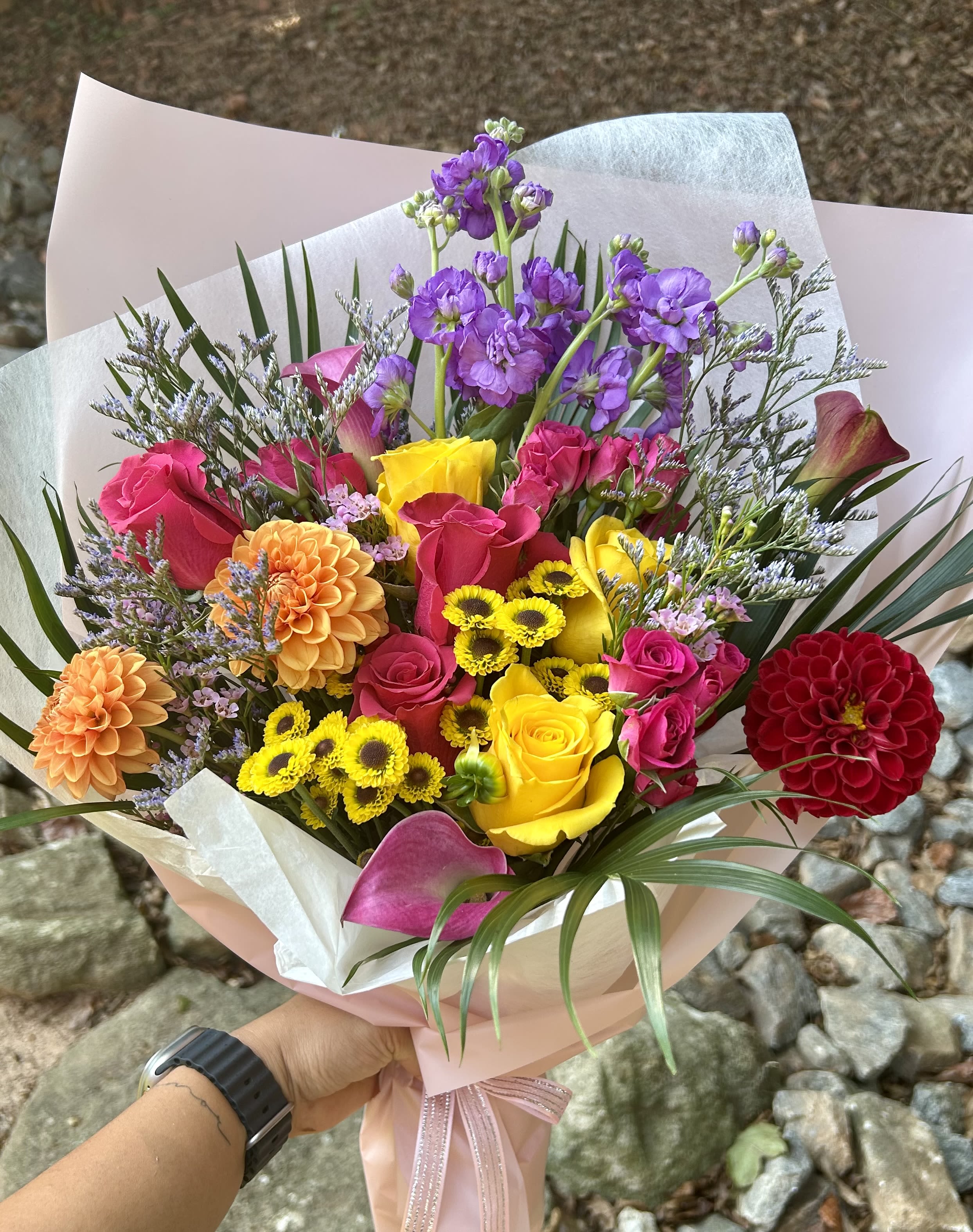 Sunset Carnival - Bursting with vibrant hues, Sunset Carnival is a joyful celebration of color and life. This wrap bouquet features radiant yellow roses, fiery orange dahlias, bold magenta calla lilies, and deep crimson zinnias—all complemented by soft purple stock, pink spray roses, and playful yellow button mums. Wrapped in delicate blush paper and tied with a satin bow, this bouquet is a perfect gift for birthdays, congratulations, or any moment worth celebrating with a splash of color.