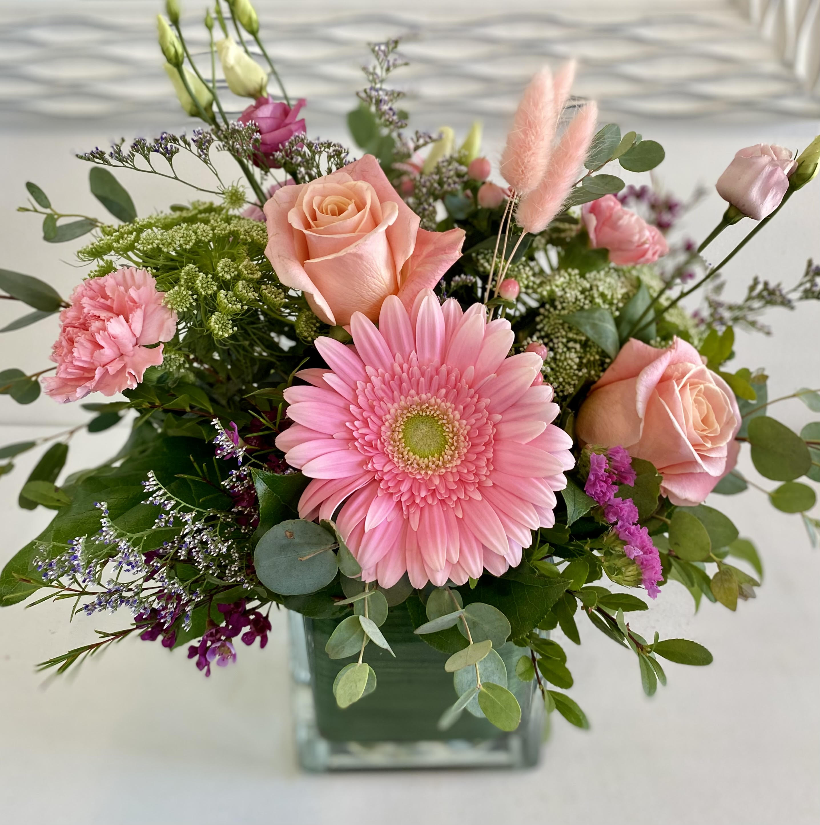 Peaches and Pinks - A charming mix of blush roses, pink gerbera daisies, carnations, and lisianthus comes together with delicate greenery and textural accents like bunny tails and filler flowers. Arranged in a modern square glass vase, this bouquet exudes elegance and sweetness—perfect for expressing love, appreciation, or celebrating a special moment.