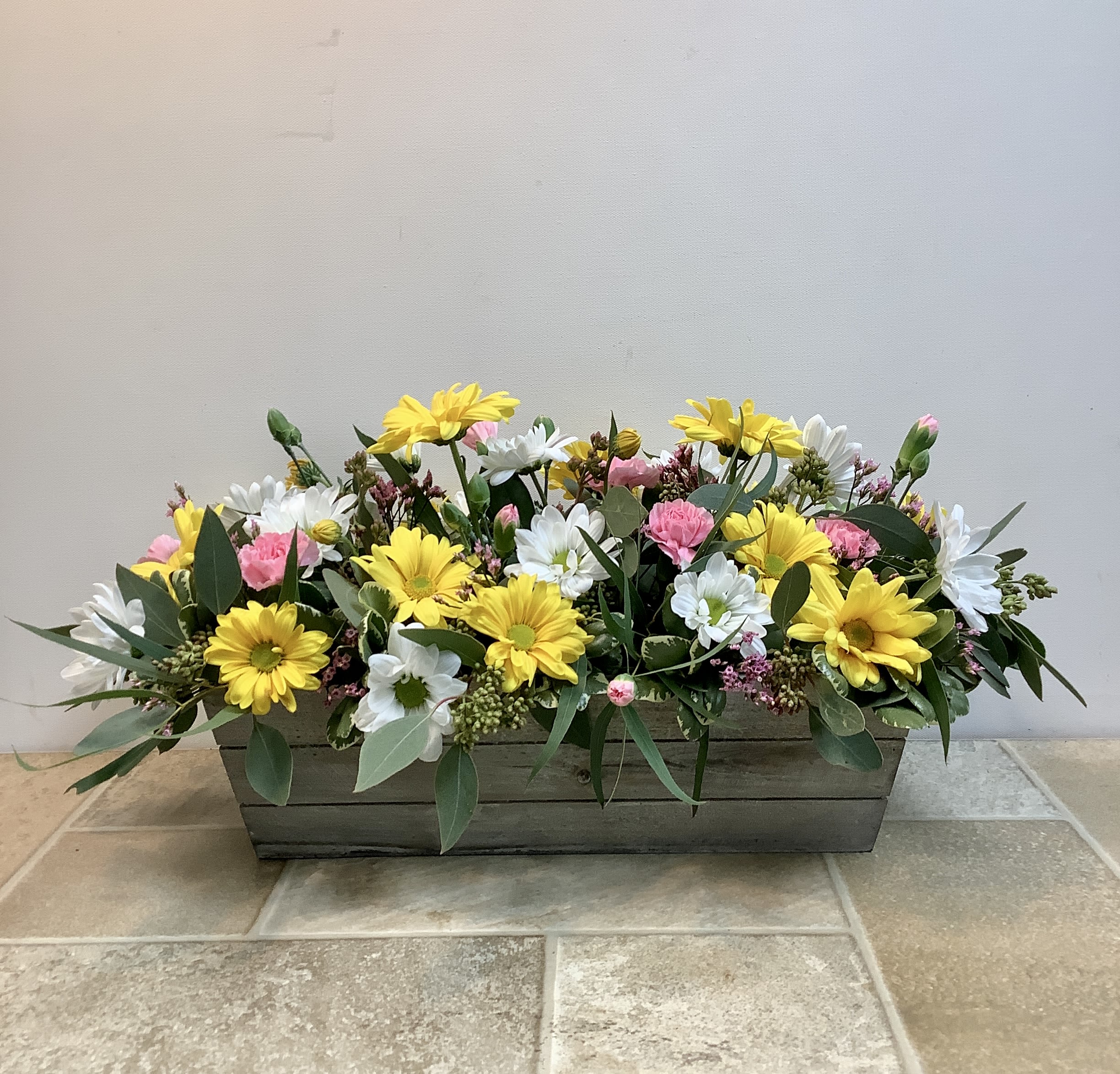 Simply Sunny - A lush, garden-style centerpiece featuring cheerful yellow daisies nestled among crisp white blooms and soft pink accents. Fresh greenery—eucalyptus and textured filler—adds movement and a natural, just-picked feel. Designed in a rustic wooden planter, the low, elongated shape makes it perfect for a dining table or entryway, bringing a warm, welcoming burst of color without blocking the view. It measures approximately 18” long x 8” high. 