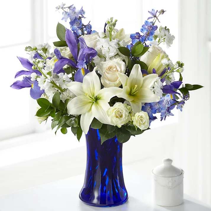 Calming Comfort Bouquet - Accented by a deep blue vase, our Calming Comfort Bouquet is crafted with iris, lilies, stock and delphinium. This unique mix of blue and white flowers makes a well-suited tribute to send to those memorializing the loss of someone who had a soothing presence.  Details The Deluxe Bouquet is approximately 19"H x 17"W. Designed by florists, ready to display. For long–lasting blooms, replace the water daily. We suggest trimming the stems every couple days.