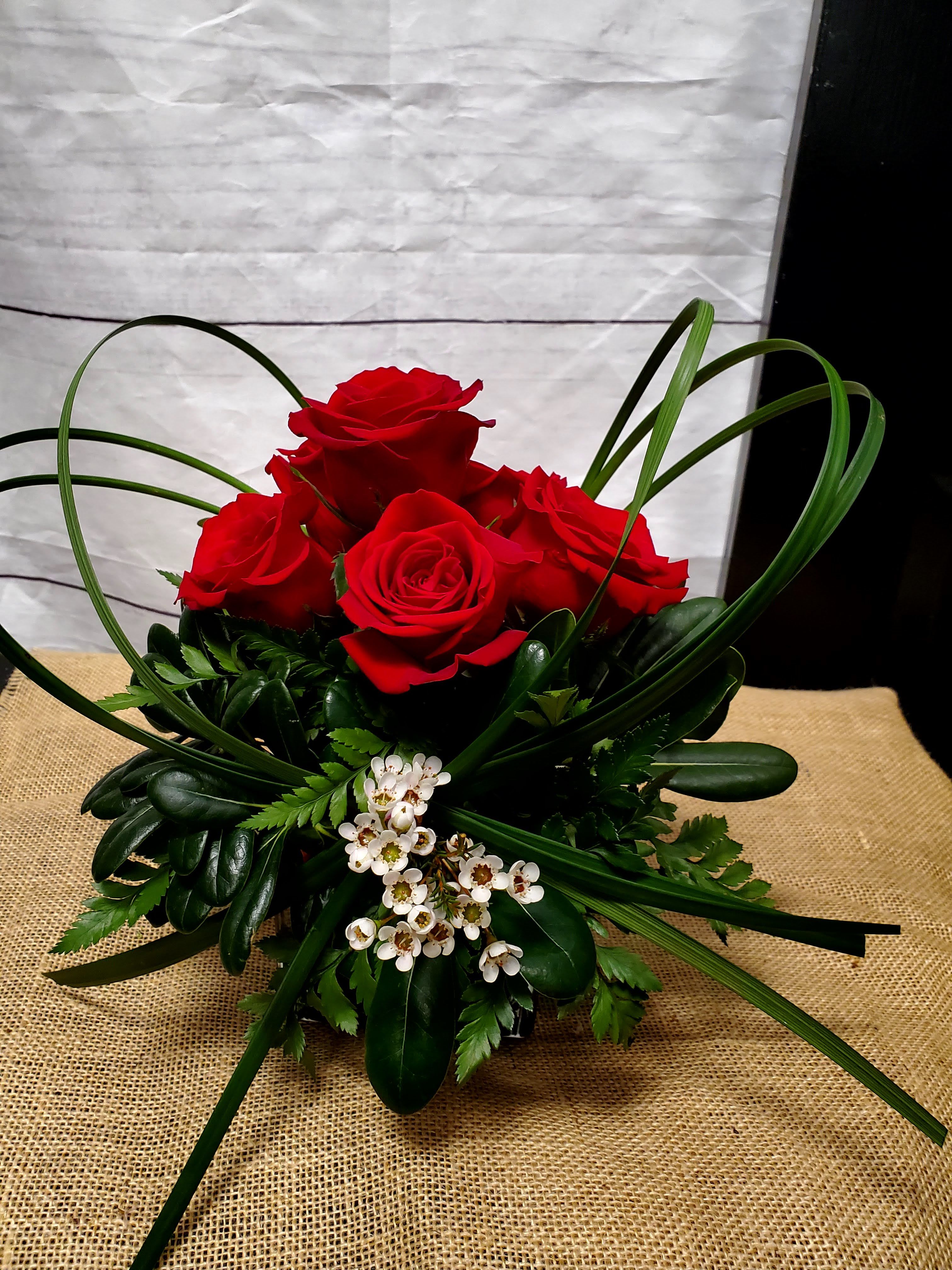 Love Cubed - This is a striking and modern floral arrangement with a bold and romantic feel.   Red roses are the focal point, symbolizing love and passion. Their vibrant color draws immediate attention. Accents of white wax flowers add a delicate contrast and a touch of elegance with their small, dainty blooms. Dark green foliage, including glossy leaves, provides a lush and structured base for the arrangement. Loops of bear grass create a dynamic and contemporary design, adding movement and flow to the arrangement. The arrangement is compact and symmetrical, making it a sophisticated centerpiece or gift for a romantic occasion. Its minimalist yet impactful style ensures it conveys a heartfelt message while maintaining a sleek aesthetic