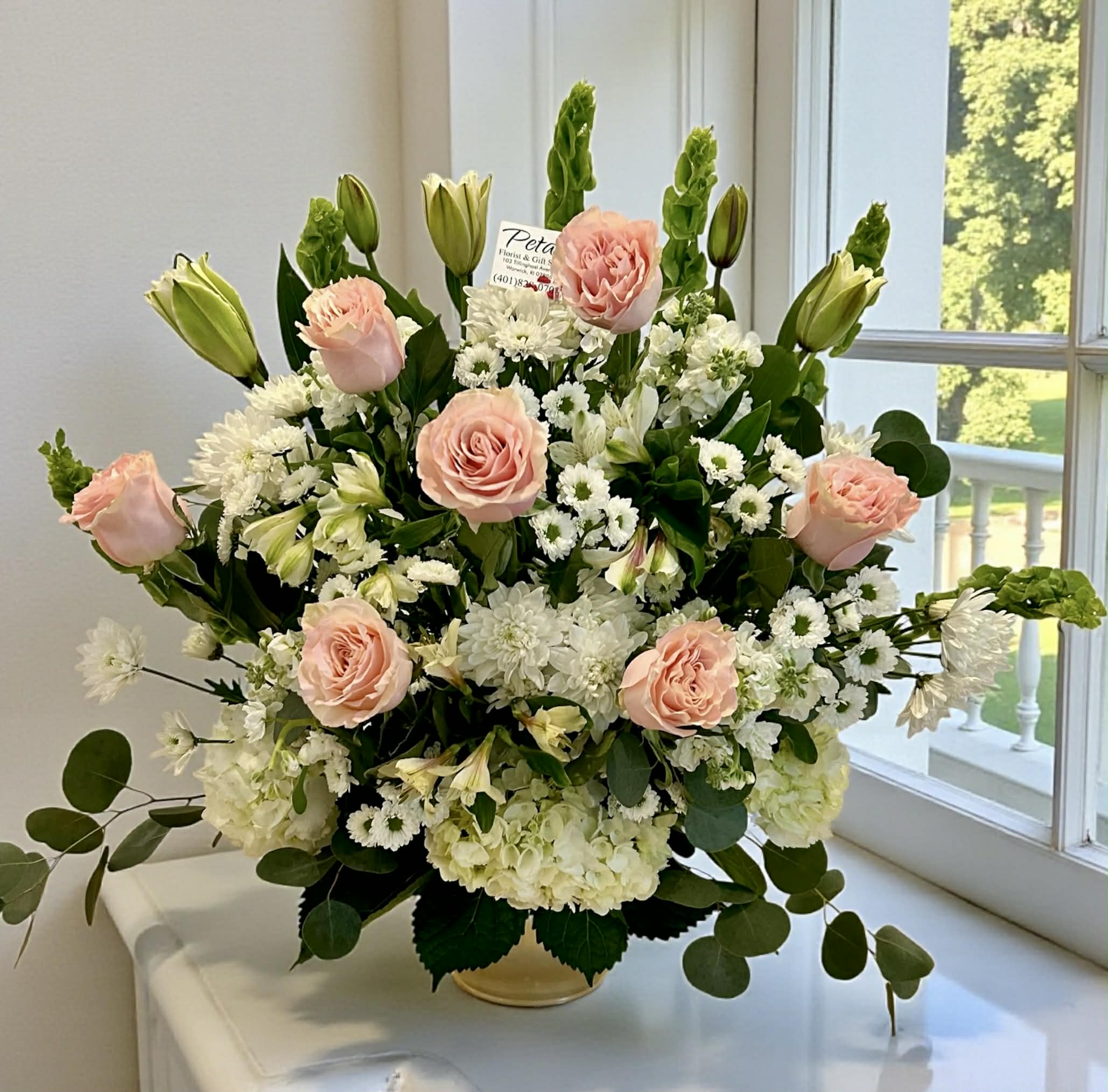Eternal Tenderness Tribute by Petals - Express heartfelt condolences and deep sympathy with this stunning white and blush pink tribute. Our "Eternal Tenderness" arrangement captures a sense of peace and honor, showcasing beautiful blush roses, white hydrangeas, elegant lilies, bells of Ireland, and chrysanthemums. This thoughtful display offers a gentle embrace during a difficult time. The soft hues of the blush roses symbolize grace, admiration, and innocent love, providing a warm, personal expression of sorrow and lasting remembrance. On occasion, some blooms may be substituted due to daily availability, but the overall aesthetic of the design will remain. 