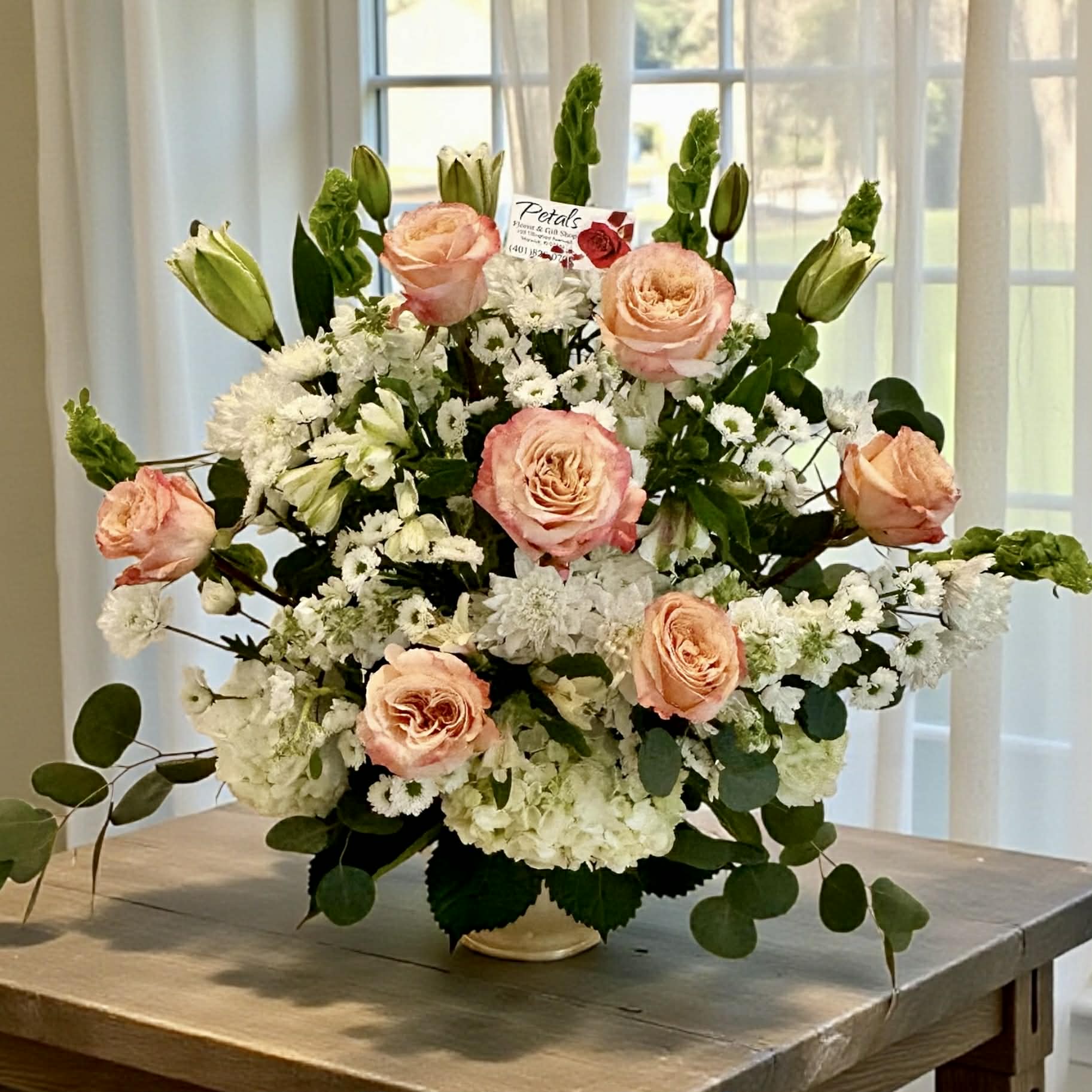 Graceful Shimmer Tribute by Petals - A graceful blend of white and peach, this Shimmer Roses tribute stands as a serene symbol of peace and reverence.  Featuring premium Shimmer Peach roses, elegant lilies, and lush hydrangeas, this forward-facing arrangement offers a warm, heartfelt embrace to those in mourning. While seasonal availability may cause slight variations, the timeless beauty and comforting aesthetic of your gift will always remain the priority.
