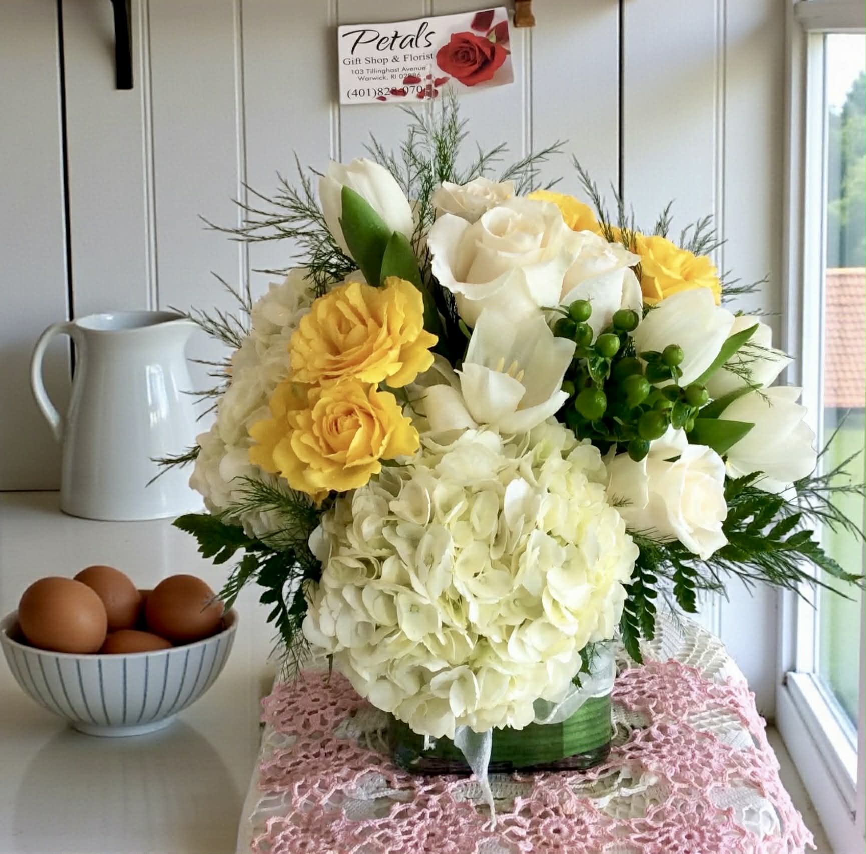 Sunny Yellow &amp; White Cube by Petals - A sophisticated yet charming arrangement featuring pristine white premium roses, sunny yellow spray roses, and fluffy white hydrangeas. The design is brightened with cheerful tulips, accented by textural hypericum berries and delicate baby's breath, all perfectly nestled among lush, delicate ferns. This professional display is artfully contained within a sleek, modern glass cube vase.