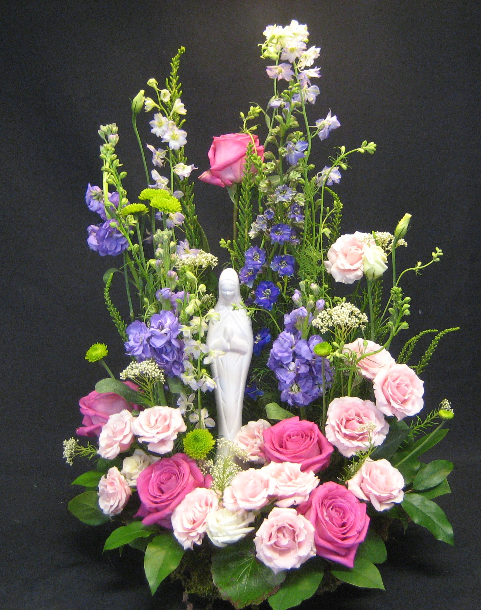 Blessed Mother Garden t16 - A white, keepsake ceramic Madonna is centered around a garden full of shades of pink roses, purple and lavender larkspur, lavender stock and more.