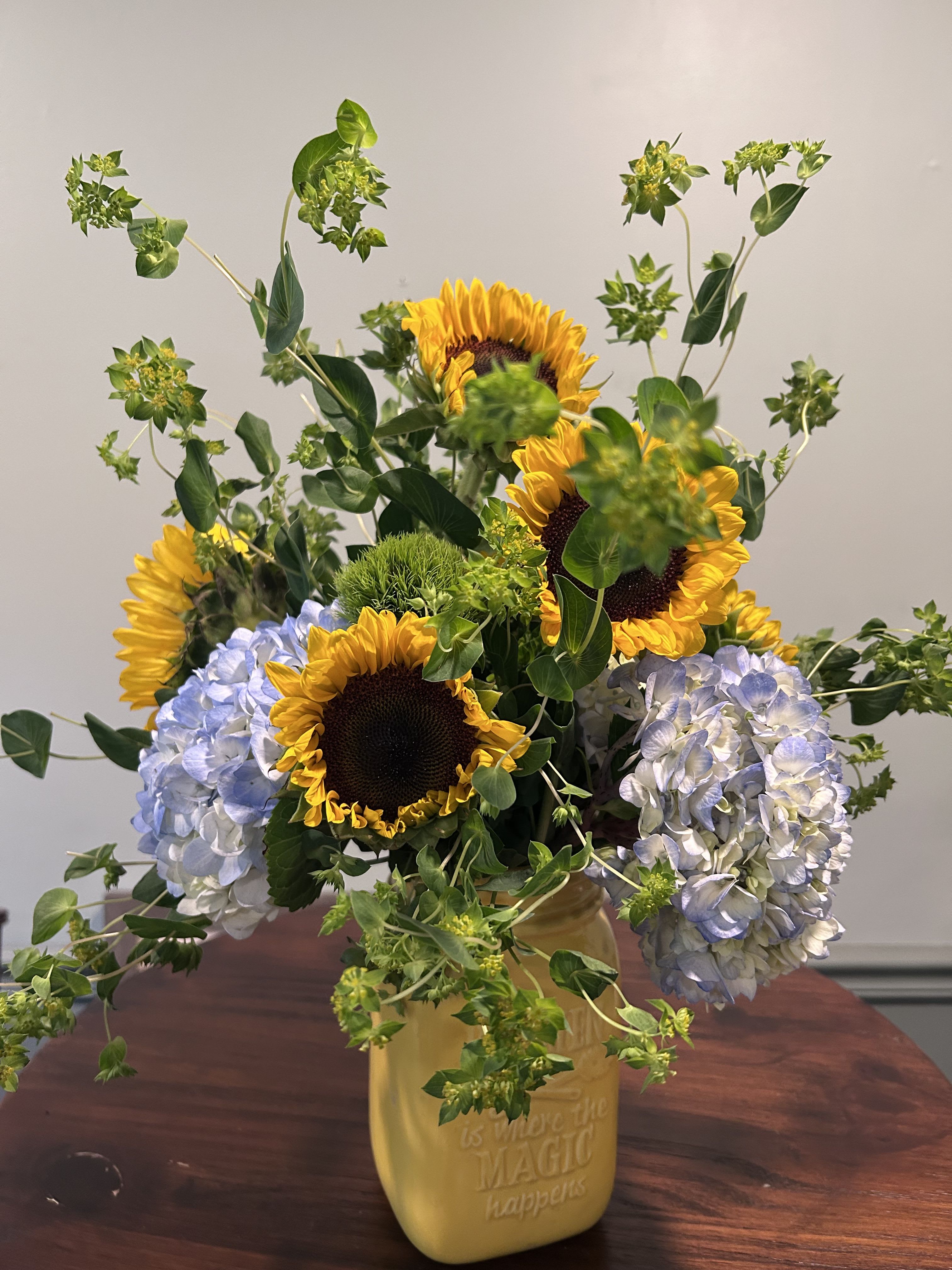 Country Charm - Add a little charm to your occasion.  An arrangement of sunflowers, blue hydrangea and buplerum has a fresh picked look. Includes a yellow mason jar.