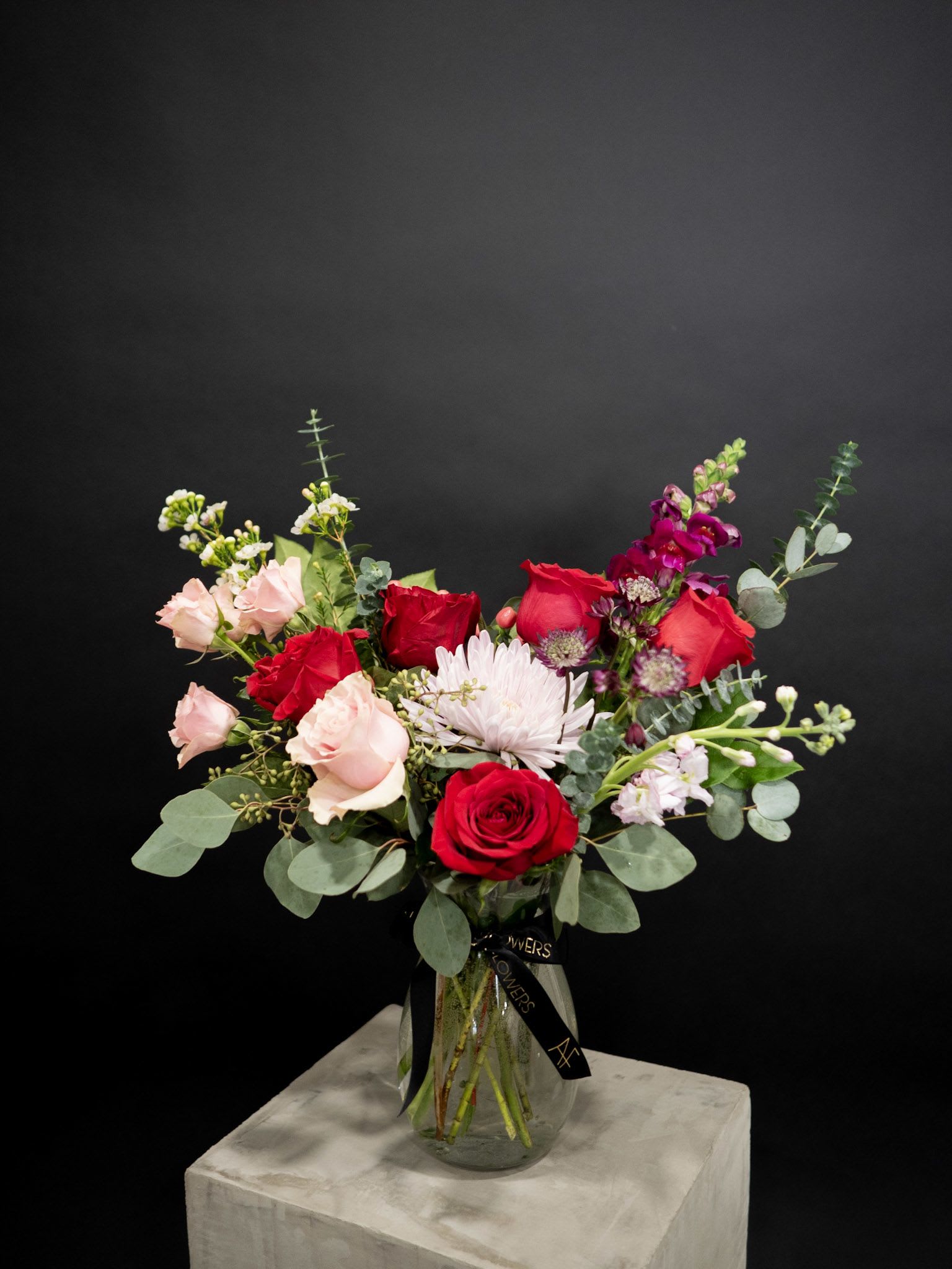 Romantic Embrace Vase Arrangement - “Romantic Embrace” captures the essence of love with its timeless beauty. This stunning arrangement features rich red roses, soft pink roses, lavender chrysanthemums, and delicate stock blooms, all nestled among lush greenery and eucalyptus. Designed in a classic glass vase, this bouquet is the perfect way to express your deepest feelings this Valentine’s Day. Vase size : 4in x 8in