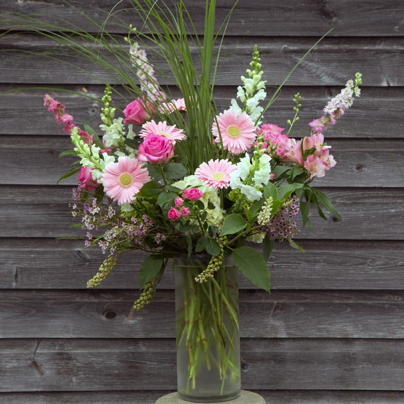 Pink Loose and Airy - Snapdragons, pink gerberas, hydrangeas, spray roses, larkspur, wax flower, rosita vendella roses and talle ornimental grass with poke weed enclosed in a a cylindrical glass vase. 