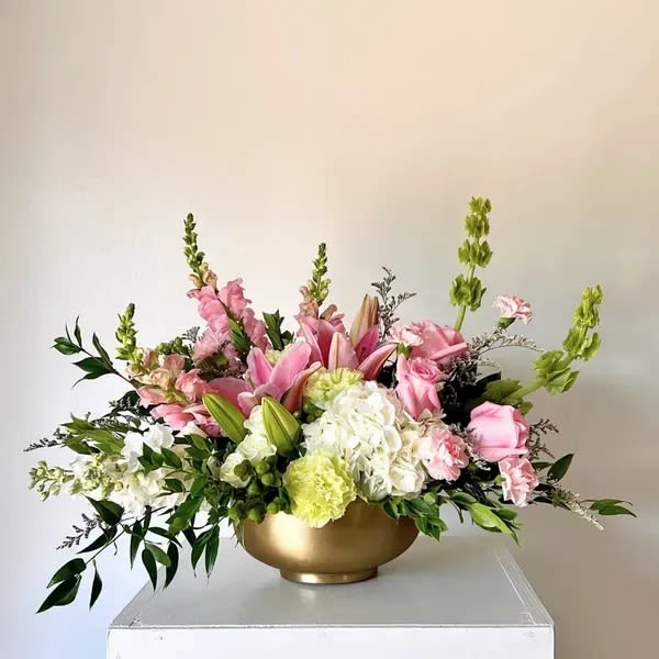 Garden Reverie - Overflowing with grace, Blush &amp; Gold Elegance features fragrant pink lilies, delicate roses, fresh hydrangea, and lush greenery designed in a radiant gold bowl. This lush, low centerpiece is perfect for weddings, special celebrations, or simply elevating a dining table with timeless sophistication.