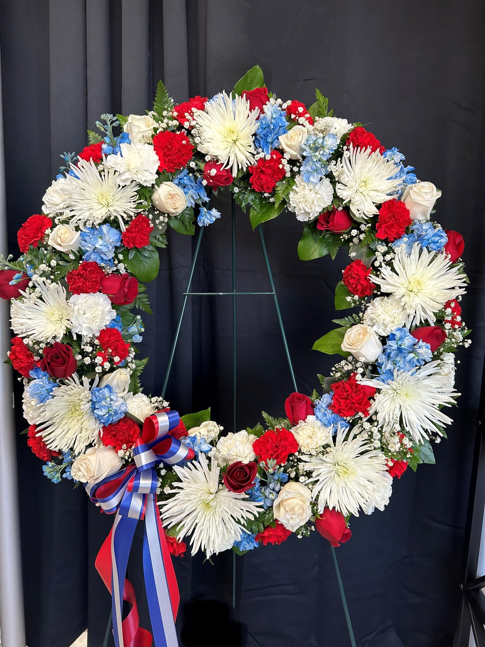 American Wreath - Standing tall in honor and remembrance, this wreath is meticulously crafted with an array of symbolic blooms. White Fuji mums and roses, red roses, and blue stock intertwine to form a poignant tribute, while white carnations add a touch of purity to the ensemble. Adorned with a tri-color bow representing the red, white, and blue of our nation's flag, this arrangement stands as a timeless symbol of patriotism and respect. May its presence offer solace and comfort to all who gather to pay their respects.