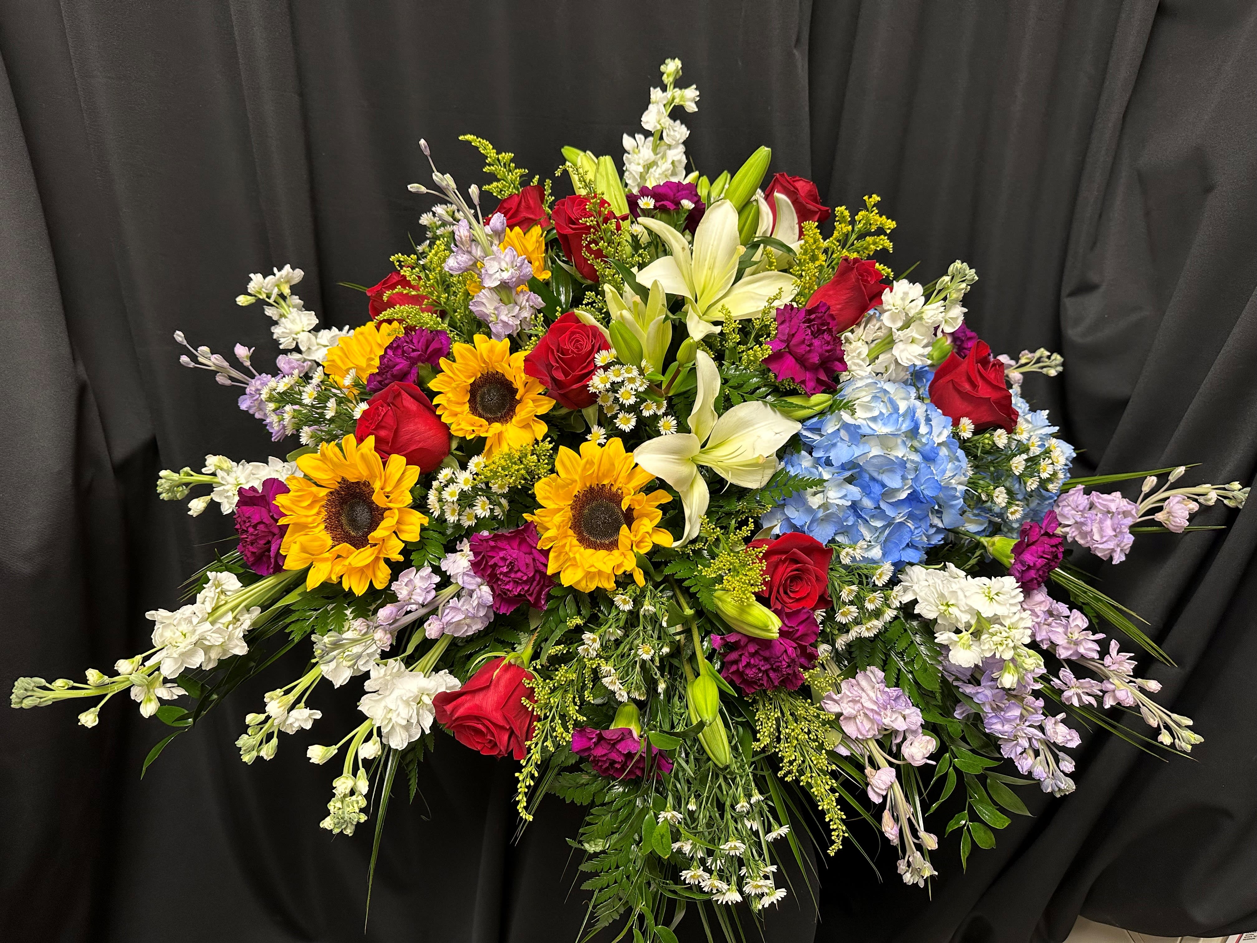 Comforting Wishes Casket Spray - Extend heartfelt wishes of comfort and solace with this half casket spray, adorned with a harmonious blend of white lilies, radiant yellow sunflowers, rich purple carnations and stock, deep red roses, and lush greenery. Each element symbolizes hope, love, and enduring memories, offering a source of warmth and joy during difficult times. May this heartfelt tribute convey your deepest sympathies and bring a sense of peace to those who mourn.