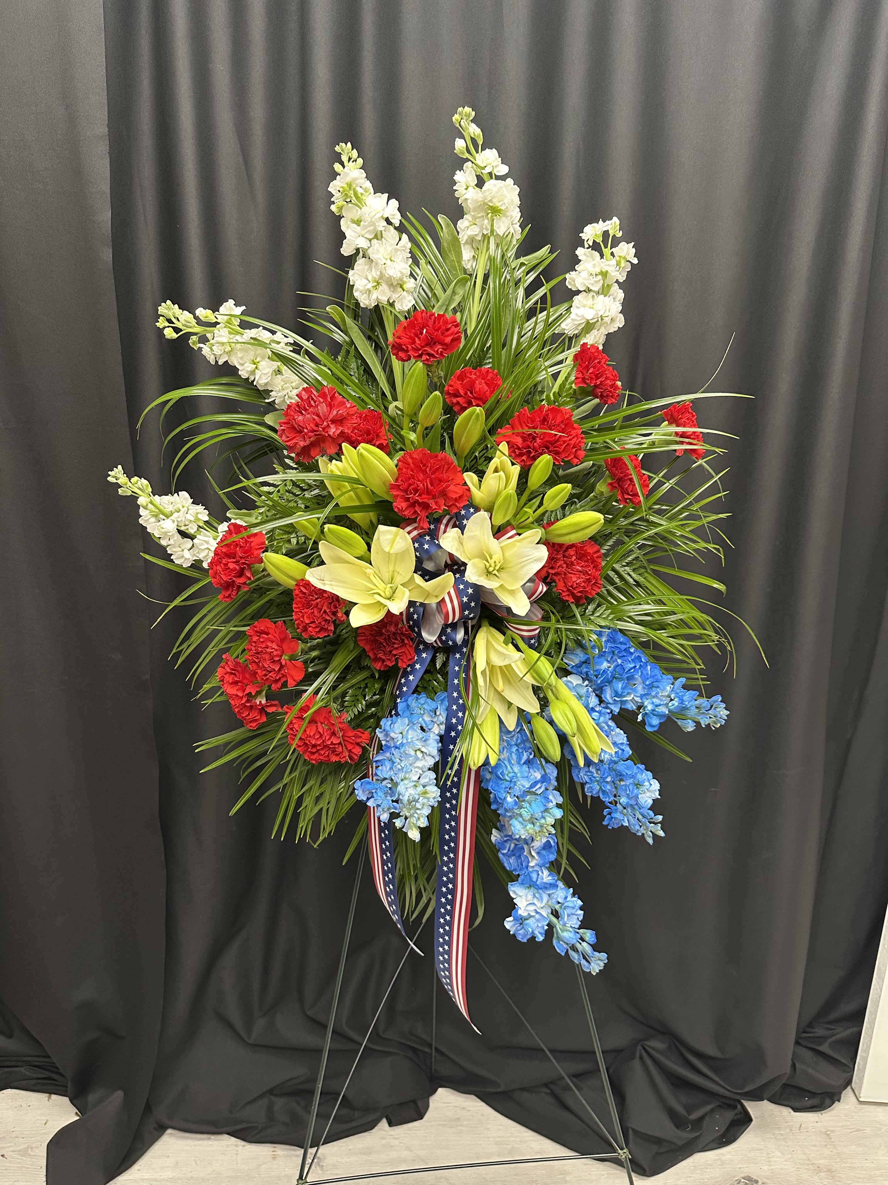 Honor and Glory - This striking standing spray is a heartfelt tribute to courage, service, and sacrifice. Featuring vivid red carnations, crisp white stock, vibrant blue delphinium, and elegant lilies, it’s accented with lush greenery and finished with a patriotic ribbon. Designed to honor the memory of a loved one who served or to commemorate national pride, this arrangement stands tall with dignity and grace.