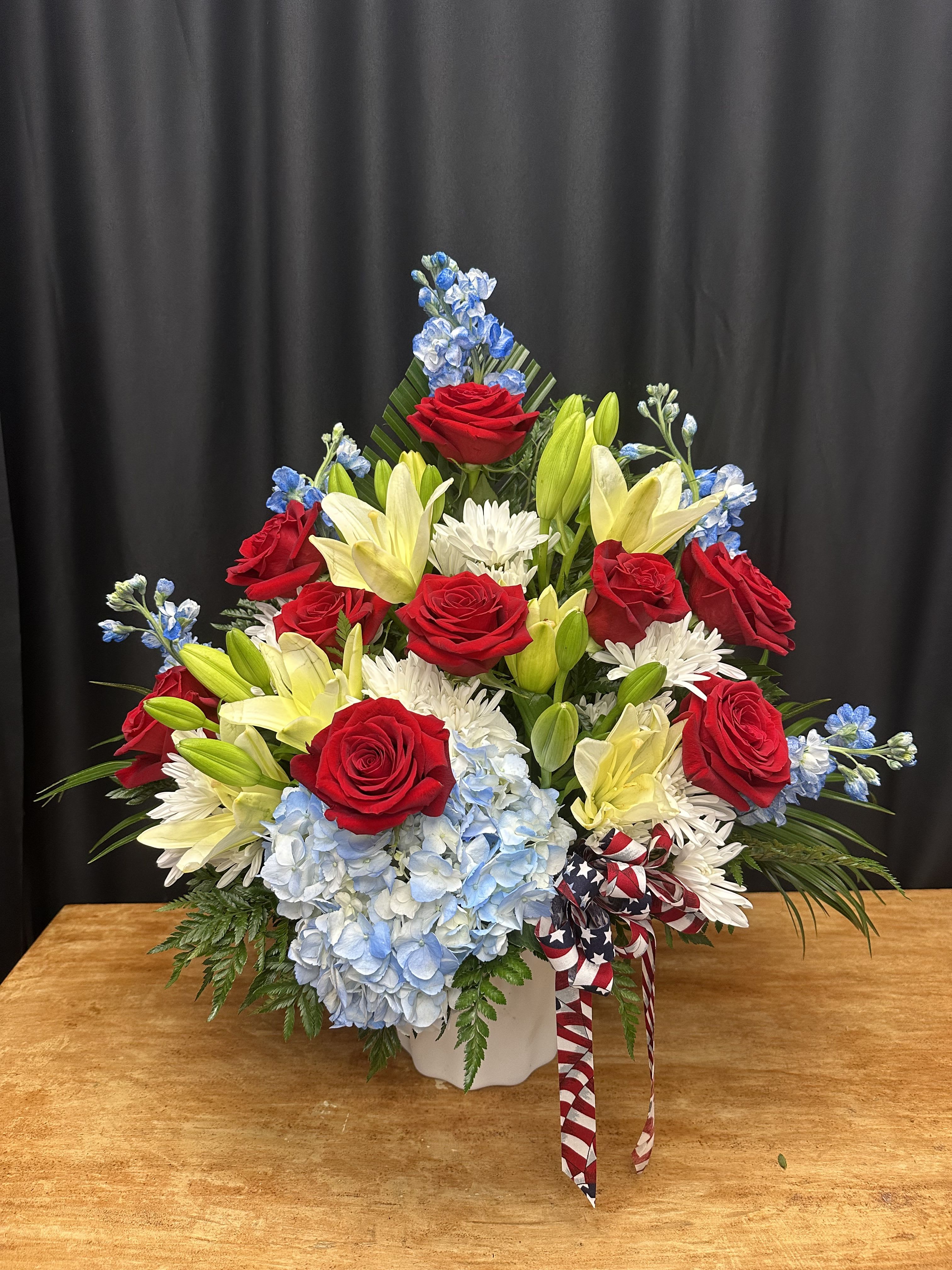Land of the Free - Celebrate American pride with this vibrant patriotic arrangement, thoughtfully designed to honor those who serve and those we hold dear. Featuring bold red roses, soft blue hydrangeas, creamy white lilies, and cheerful white mums, this arrangement is accented with blue delphinium and a stars-and-stripes ribbon. Perfect for Memorial Day, Independence Day, Veterans Day, or as a heartfelt tribute to a loved one.  Presented in a classic white container, this design is both elegant and uplifting — a true expression of freedom, gratitude, and remembrance.