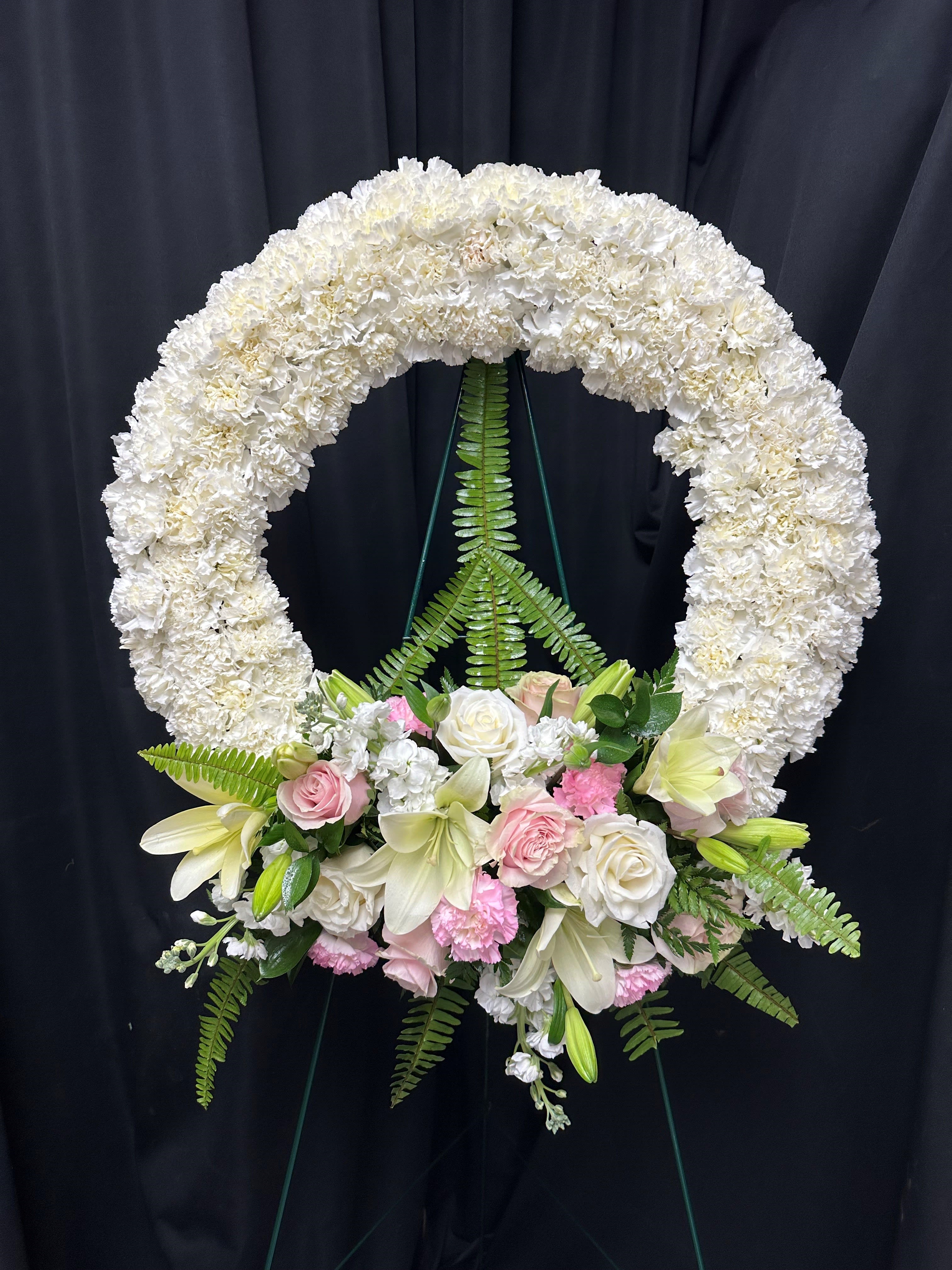 Peace and Love - This wreath of delicate white mums exudes a serene aura, accompanied by the verdant embrace of green ferns, the purity of white lilies, the soft elegance of pink roses, and the tender beauty of pink carnations. Together, these blooms create a tranquil tableau, offering solace and comfort in times of remembrance. May the peaceful presence of this arrangement bring gentle reassurance to those who mourn, enveloping them in love and warmth.    