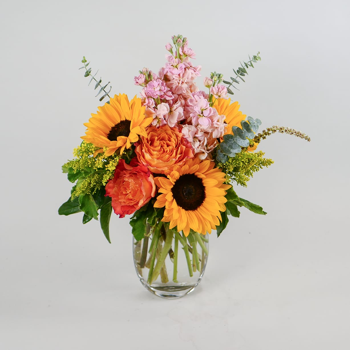 California Sunset - This bright arrangement displays a fun collection of flowers, including sunflowers, roses, fragrant stock, solidaster and other greenery in a medium glass vase. Arrangement approximately measures 17 inches tall and 12 inches wide.