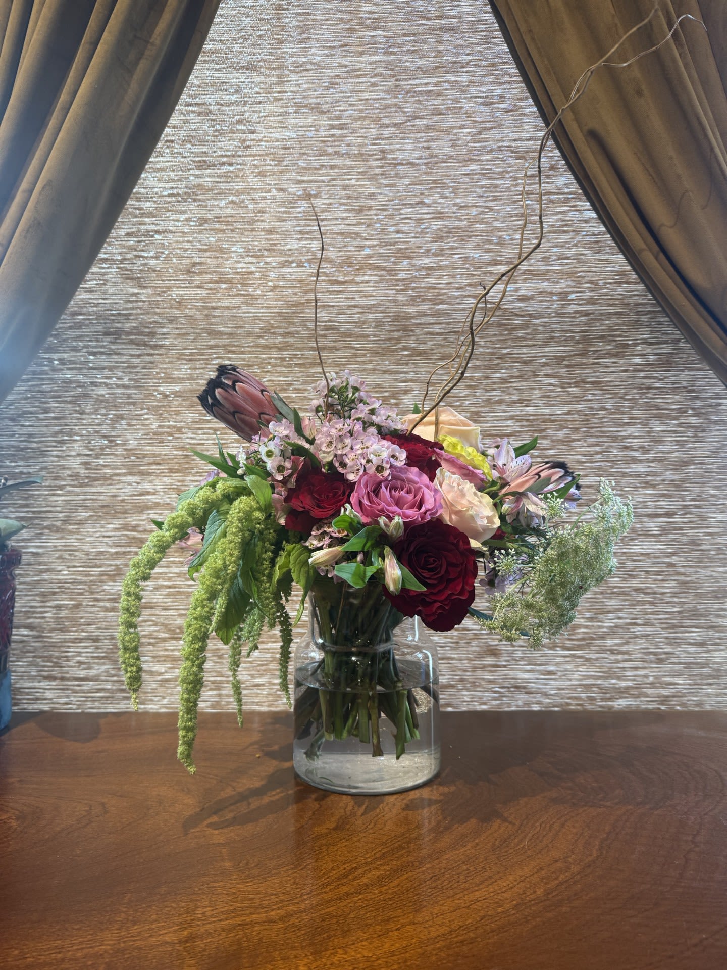 Midnight Moment Arrangement - A dramatic, high-impact statement designed to stop time.  Our most elevated Valentine’s arrangement, Midnight Moment showcases premium garden roses—Queen of Hearts, Queen’s Crown, and Phoenix—selected at peak openness for richness, depth, and intensity. Sculptural protea anchors the composition with bold structure and an editorial edge, while elegant branches create a cinematic silhouette and a sense of motion above the blooms. Luxe, confident, and unforgettable—made for grand gestures and after-dark romance.