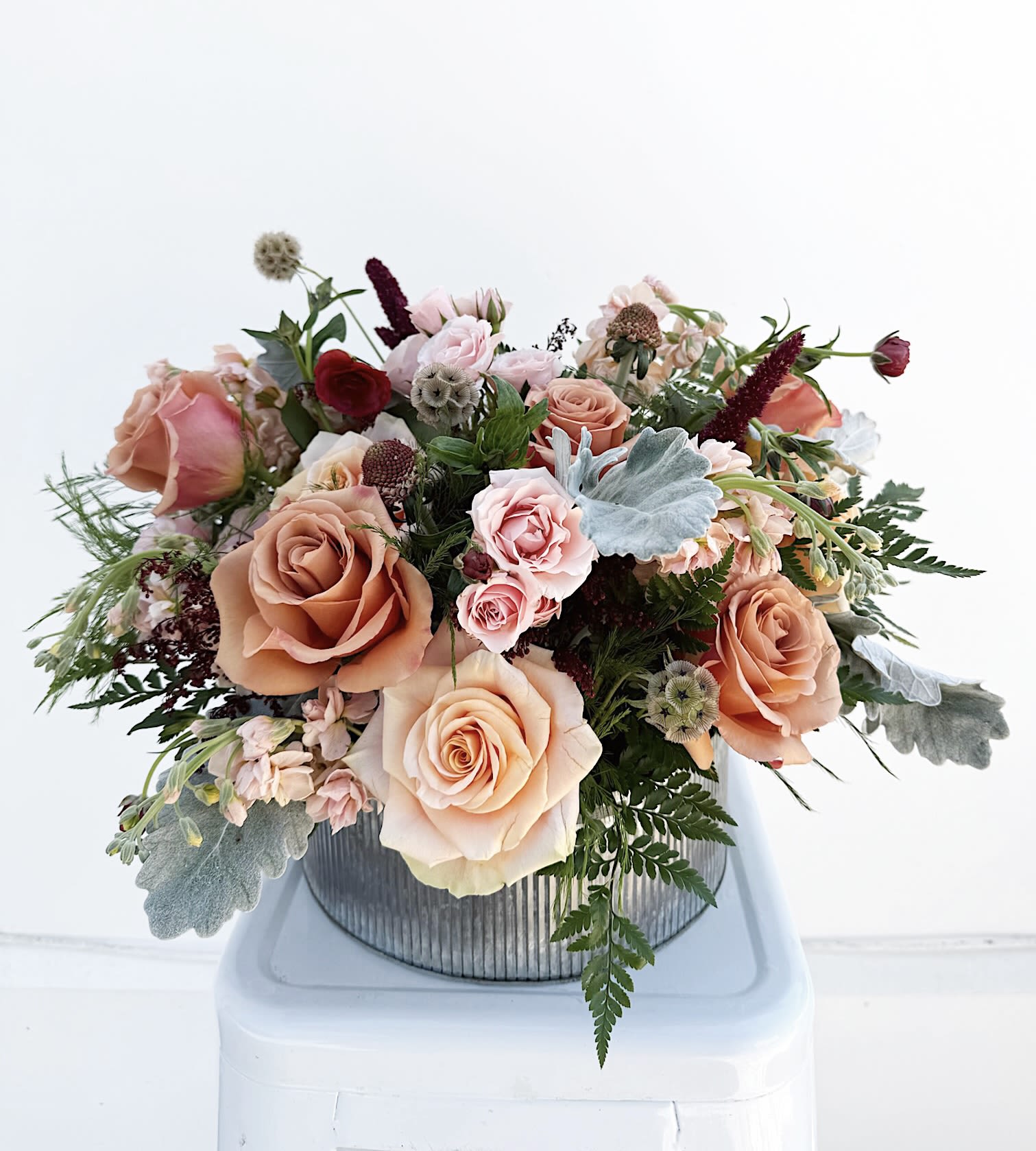 Sweet Ophelia - A charming, medium-sized arrangement bursting with sweet blooms and lush greenery in soft blush, rich burgundy, and warm peach tones. Each arrangement is thoughtfully crafted and may feature a mix of spray roses, garden roses, ranunculus, wildflowers, and other seasonal favorites, creating a naturally beautiful, romantic display. If necessary, the vase may vary due to availability but will remain similar in style.   **Please note: The deluxe arrangement is pictured***