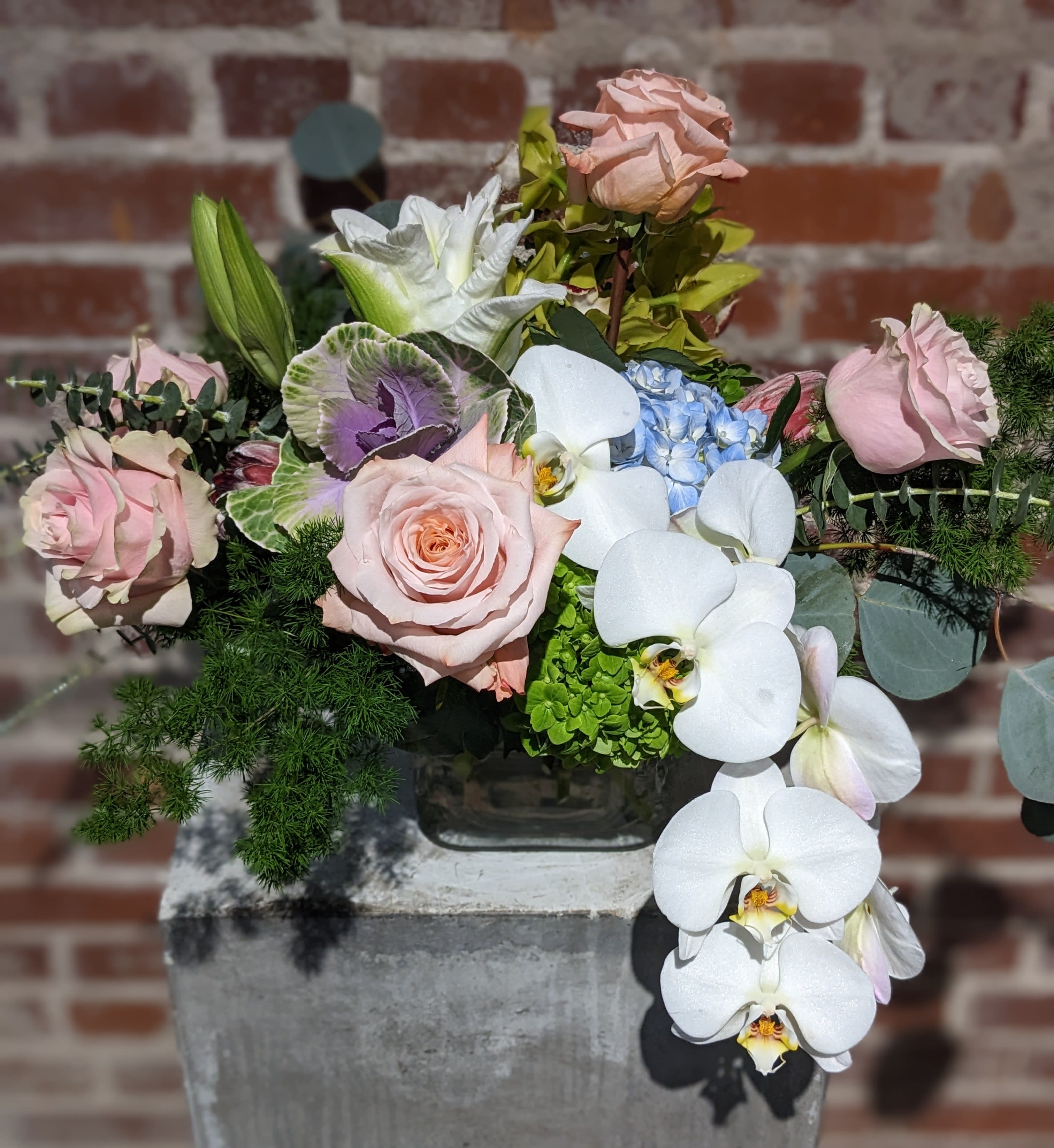 Satene - Soft pastel floral mix in a variety of texture and depth accented with rich foliage. Presented in a clear glass container with exposed stems.