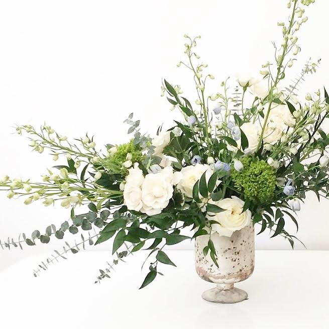 The Charlotte - A dreamy, garden-inspired arrangement brimming with airy whites and lush greenery. It may include spray roses, garden roses, stock, delphinium, eucalyptus, ruscus, and other seasonal blooms, all elegantly arranged in a marbled mercury glass pedestal vase. Perfect for adding effortless sophistication to any setting.