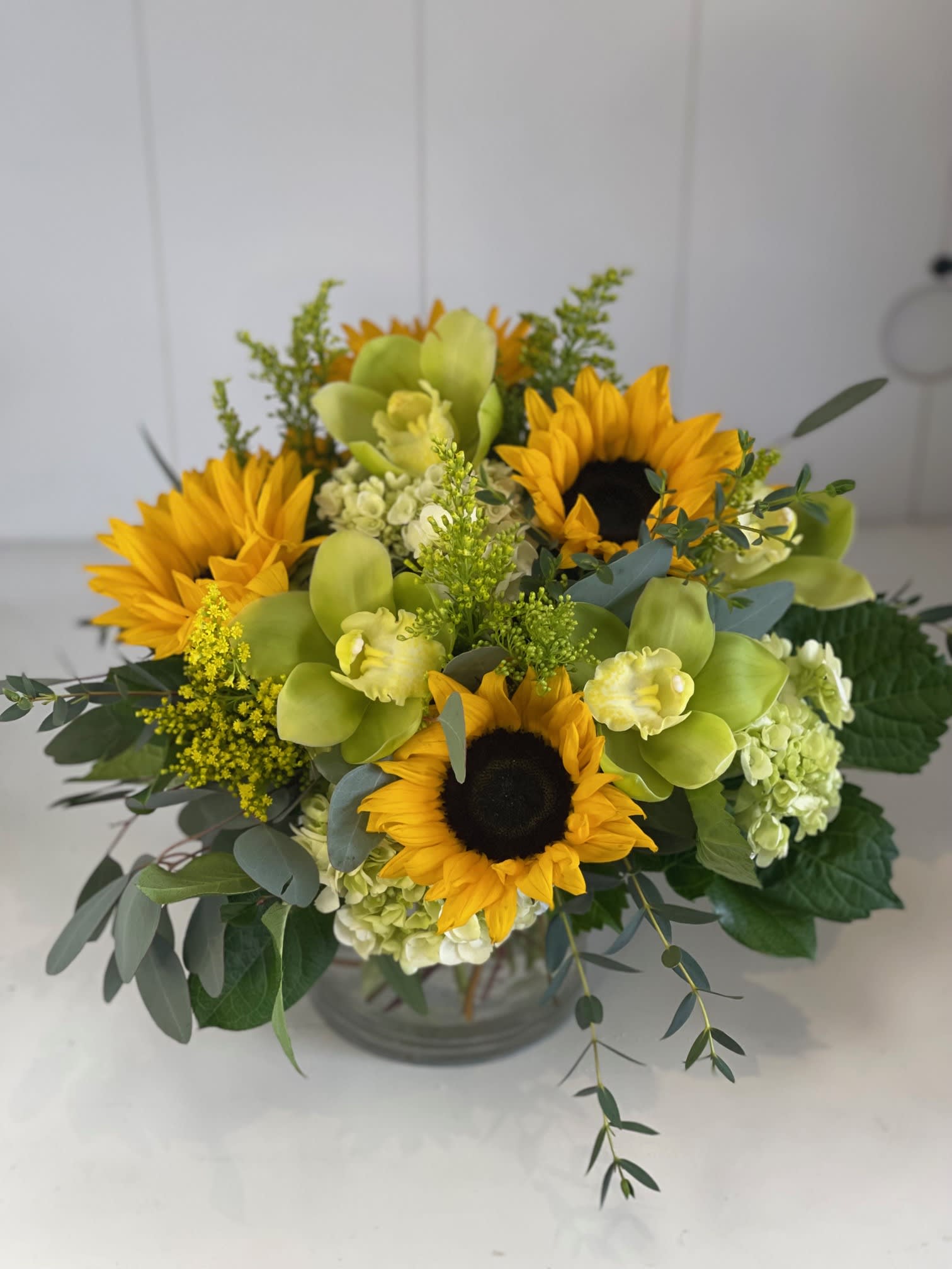 Spring Harvest  - A beautiful combination of golden sunflowers, mini green hydrangeas, and exotic cymbidium orchids, complemented by a touch of rustic greenery. This arrangement exudes warmth and charm, perfect for adding a touch of nature's beauty to any occasion, whether it's a celebration or a simple moment of joy.