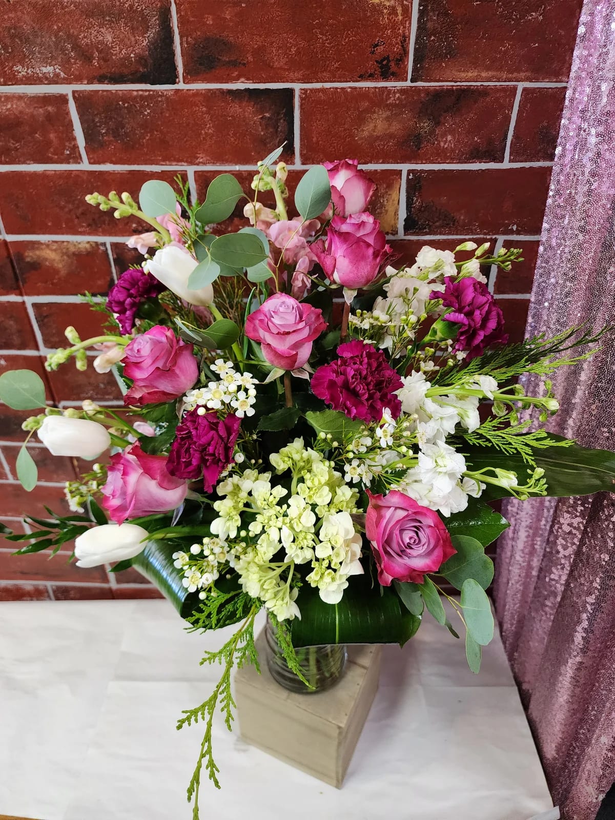 Isn't She Lovely - This one sided design has "deep purple" roses, White Tulips, white stock, light pink snap dragons, Green hydrangea, carnations, and a mix of greens in a vase.  * Some stages of flower life may vary. (Example: Lilies may be more open than imaged.) Flower shortages and product availability means that some substitutions will apply. Value will always be filled. This bouquet might include eucalyptus amongst the greenery. It may be toxic to pets including but not limited to Cats and Dogs if digested.