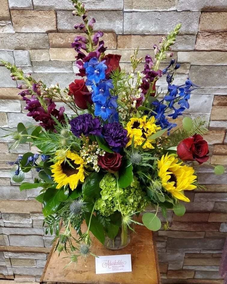 Jewel Toned Garden - A beautiful one side bouquet, designed with fresh flowers to include sunflowers, roses, delphinium, snapdragons thistle, and carnations.  * Some stages of flower life may vary. (Example: Lilies may be more open than imaged.) Flower shortages and product availability means that some substitutions will apply. Value will always be filled.  This bouquet might include eucalyptus amongst the greenery. It may be toxic to pets including but not limited to Cats and Dogs if digested.