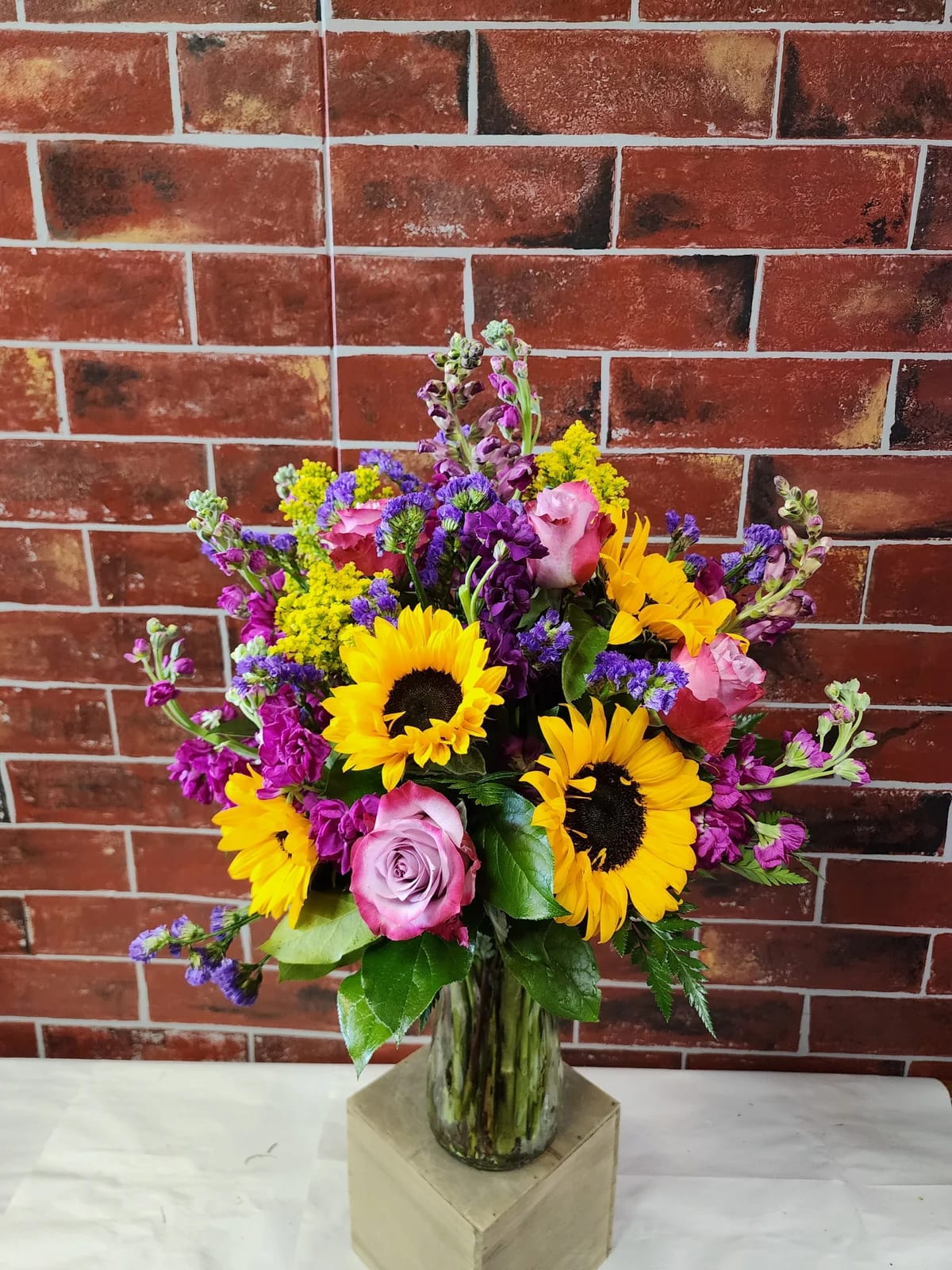 Just for You - This one sided design has "deep purple" roses, pink and purple stock, purple snapdragons, sunflowers, and a mix of greens in a vase.  * Some stages of flower life may vary. (Example: Lilies may be more open than imaged.) Flower shortages and product availability means that some substitutions will apply. Value will always be filled. This bouquet might include eucalyptus amongst the greenery. It may be toxic to pets including but not limited to Cats and Dogs if digested.