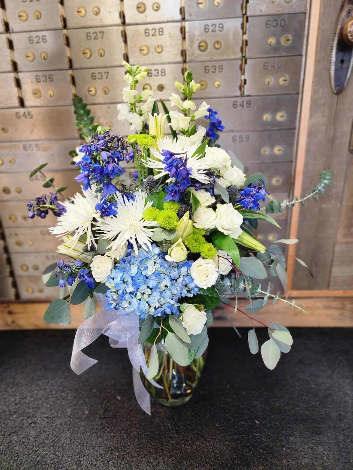 Sincerely Designed - A one-sided flower arrangement designed with fresh flowers to include blue delphinium, blue Hydrangea, white spider mums, green button, white spray roses, white lilies and white snap dragons in a vase with a bow.  * Some stages of flower life may vary. (Example: Lilies may be more open than imaged.) Flower shortages and product availability means that some substitutions will apply. Value will always be filled. This bouquet might include eucalyptus amongst the greenery. It may be toxic to pets including but not limited to Cats and Dogs if digested  Designer: Susan Rademaker