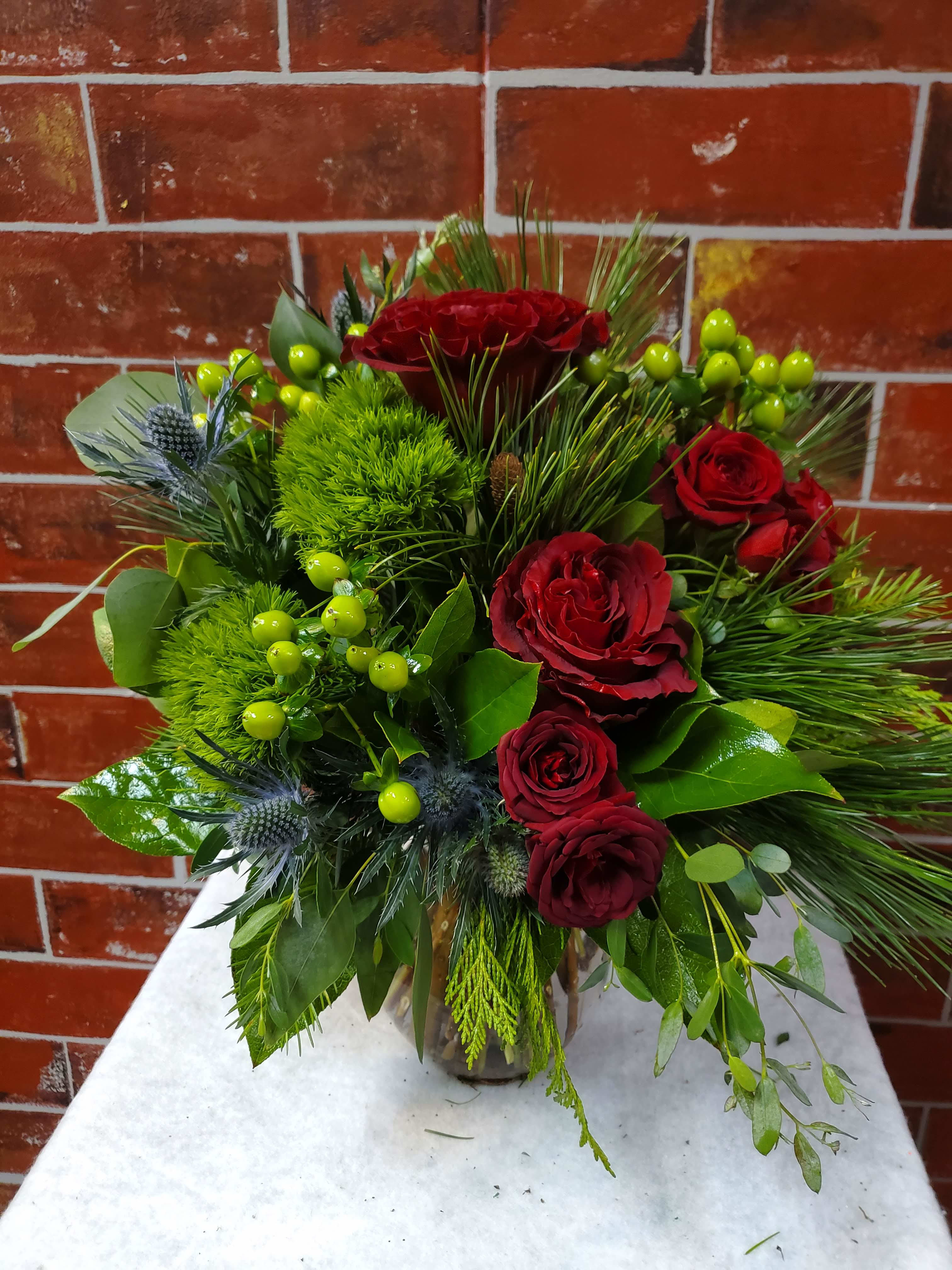 Winter Love Story - Red roses, green hypericum berries, blue thistle, green Trix, designed with winter greens in a vase.  *Some stages of flower life may vary. (Example: Lilies may be more open than imaged.)  Flower shortages and product availability means that some substitutions will apply. Value will always be filled.  This bouquet might include eucalyptus amongst the greenery. It may be toxic to pets including but not limited to Cats and Dogs if digested.