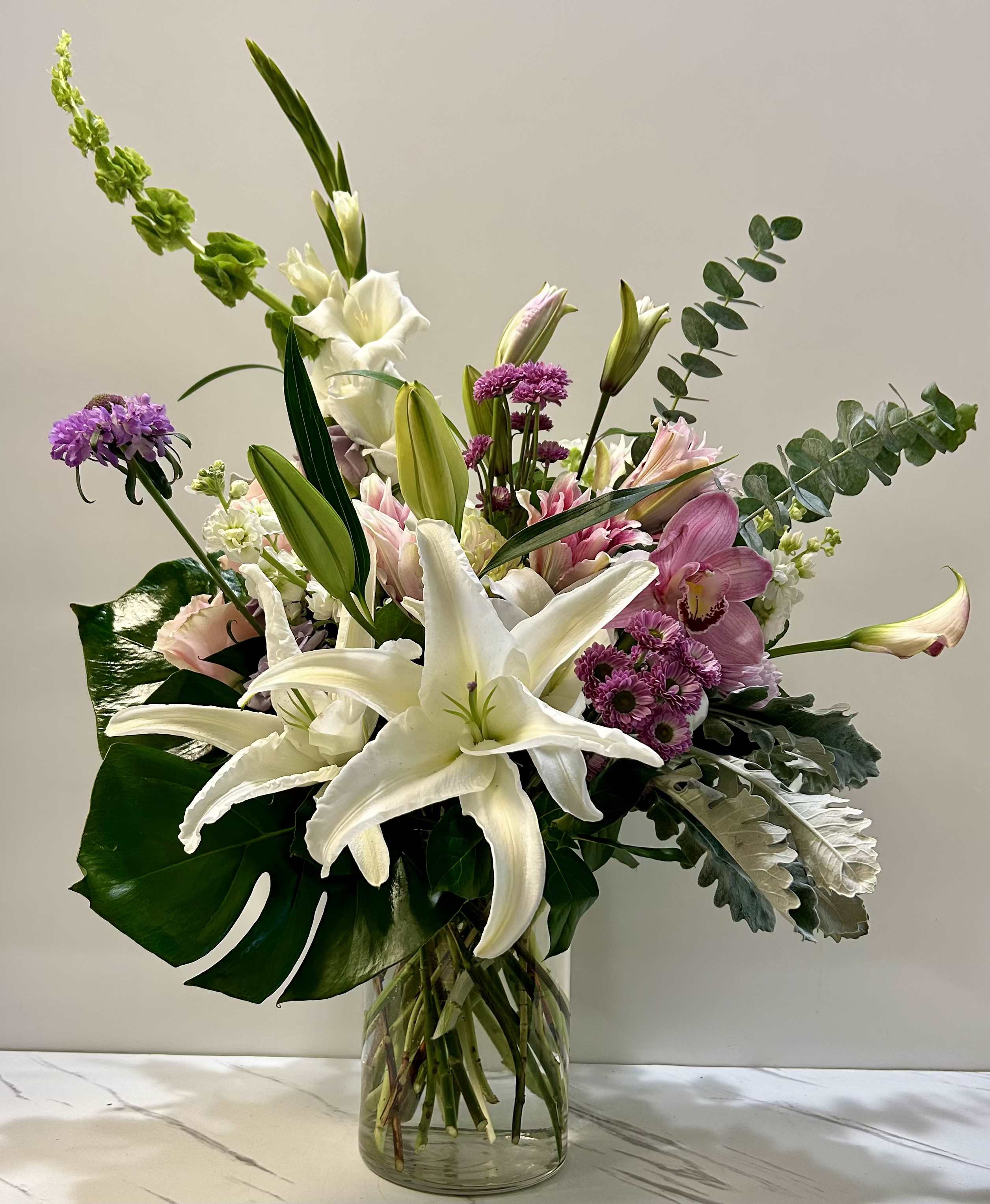 Jardin de Lumière - A breathtaking composition of pristine lilies, delicate pinks, and soft lavenders, artfully arranged to capture the essence of a radiant spring garden. This refined design exudes elegance and grace, evoking the serenity and light of a blooming paradise. Approximate size: 15" x 20".