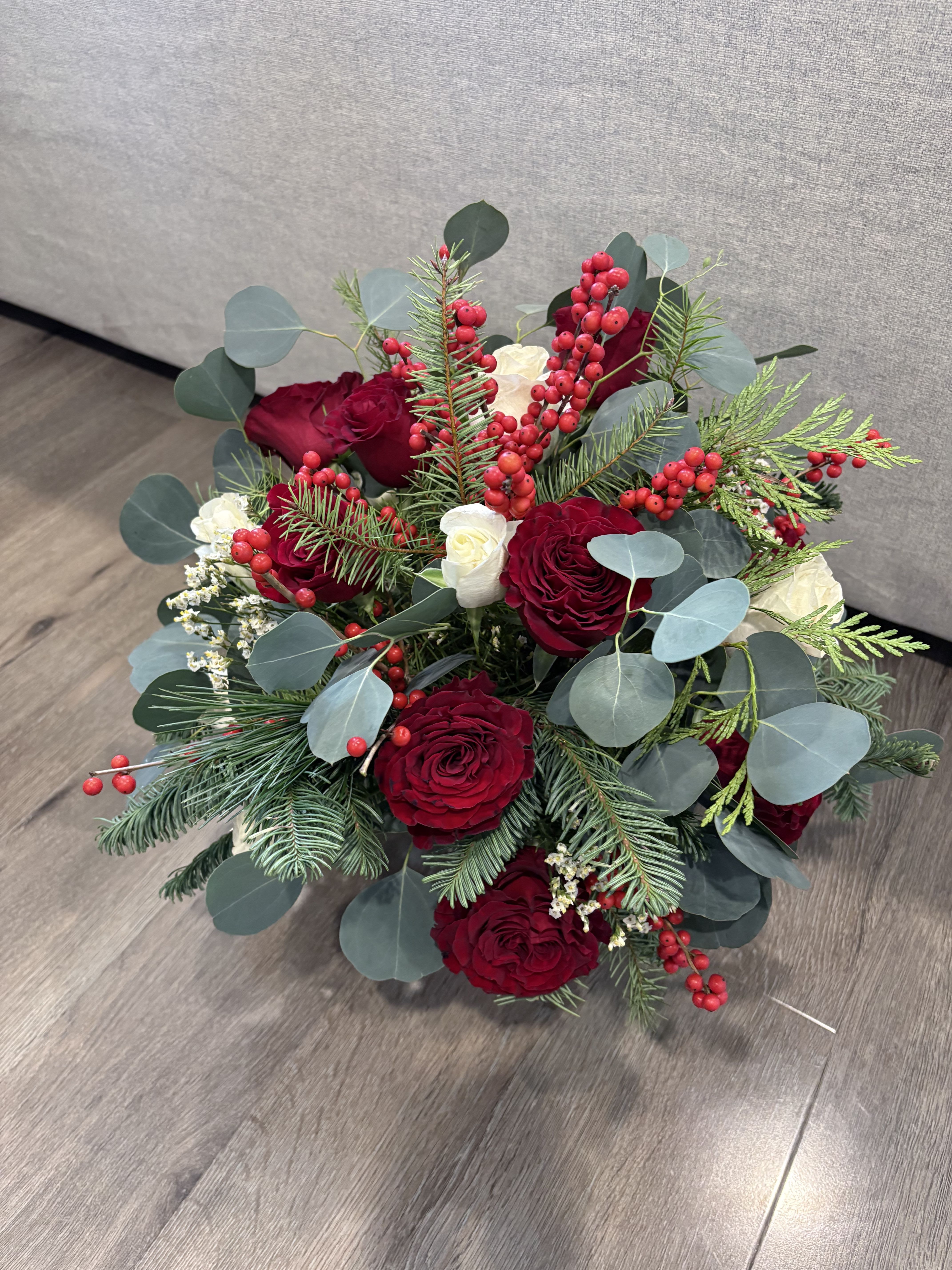 Elegant Festive Floral Arrangement - This beautifully arranged bouquet features vibrant red roses and white roses, surrounded by lush greenery, including eucalyptus leaves and assorted foliage. Sprigs of pine and red berries add a festive touch. 
