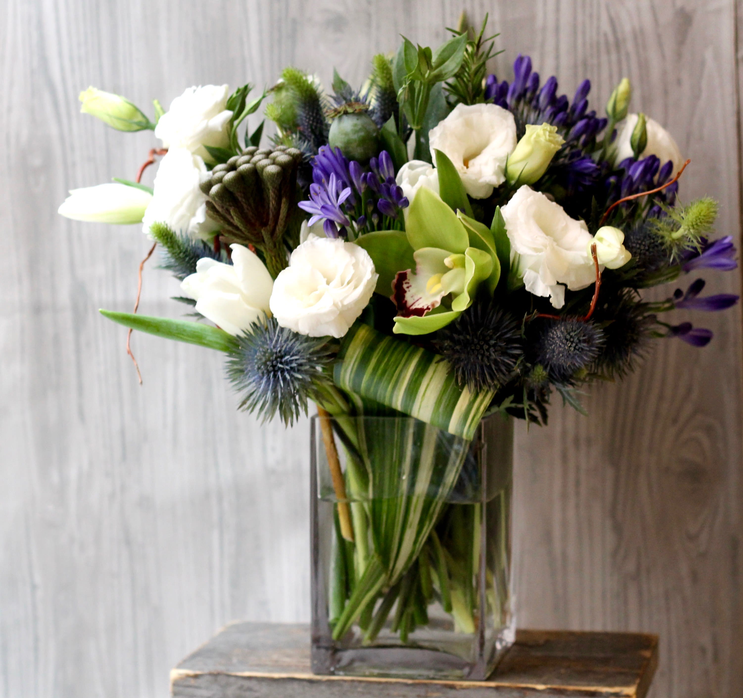 Blue Thistle  - Crisp white and green flowers with blue thistle accent. Designed in clear 5x3 vase, a clean compact design perfect for a desk. Upgrades will add more premium flowers but the size will not change very much.