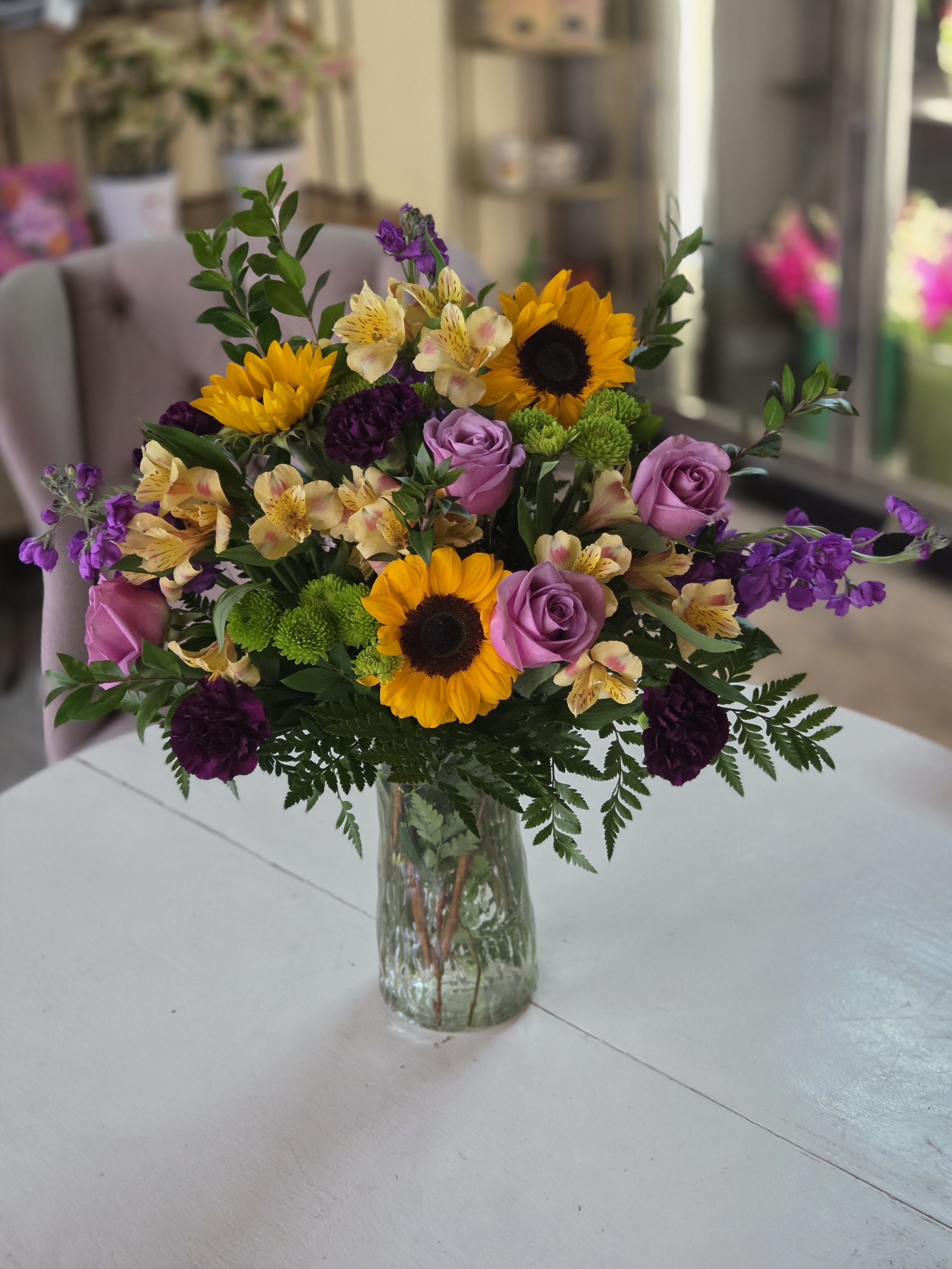 Honey Glow - Combining vibrant sunflowers with soft lavender roses, buttery alstroemeria, deep plum carnations, and bright green button mums, this arrangement would be perfect as a joyful celebration piece or an uplifting gift, it balances bold color with refined texture for an elegant, sunlit statement.