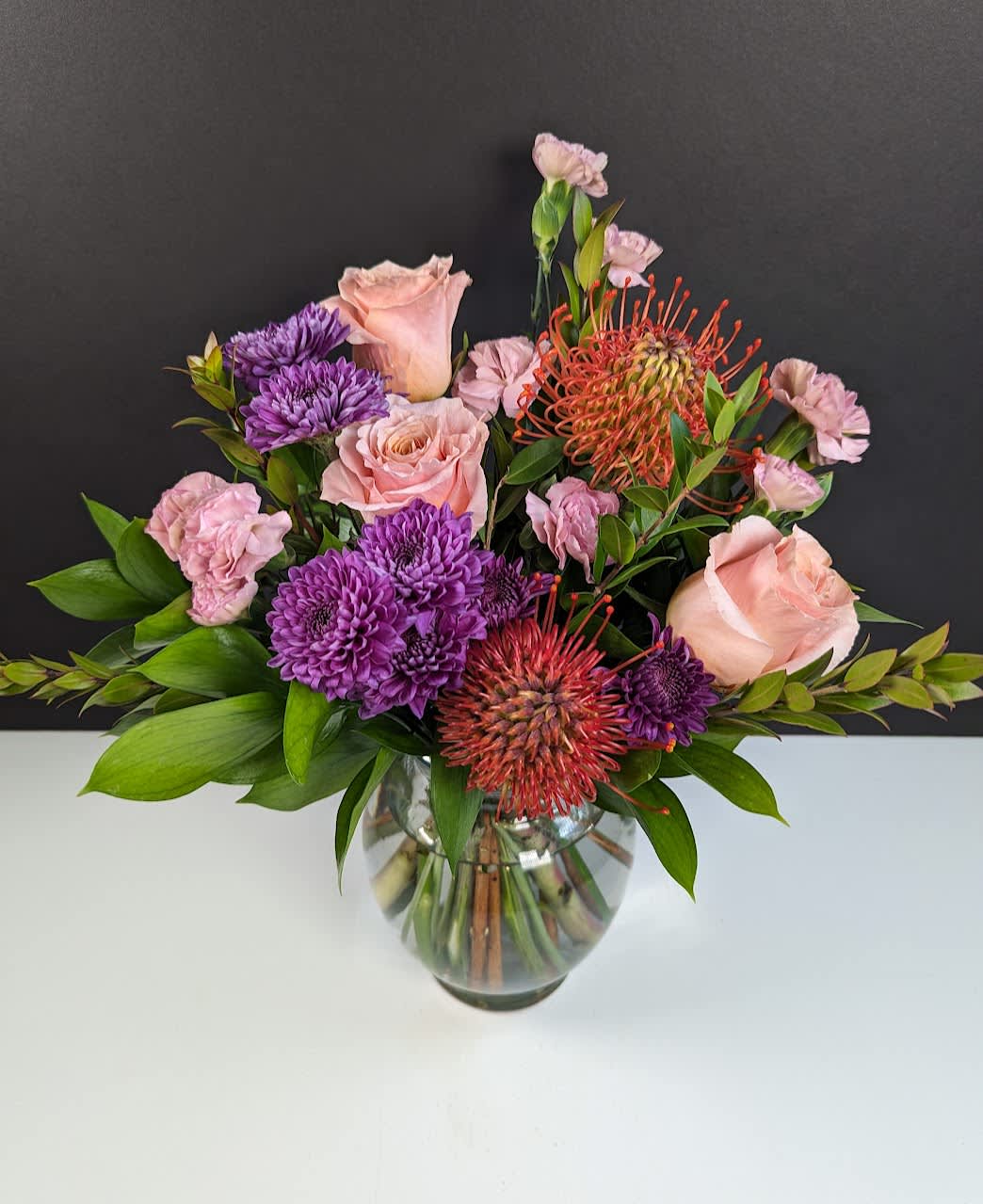 Sunset Bliss Floral Arrangement  - Experience Serene Luxury with the Sunset Bliss Floral Arrangement  Elevate your space with Sunset Bliss, a masterpiece of sophistication and natural beauty inspired by the warm, vibrant hues of a setting sun. This premium arrangement features:  -Exquisite orange pincushion protea for a bold, sculptural focal point -Luxurious pink roses, hand-selected for their lush blooms -Rich purple spray mums, adding depth and texture -Delicate mauve mini carnations, a soft touch of elegance  Perfect for creating an ambiance of refined tranquility, Sunset Bliss is more than a floral arrangement—it's an experience of timeless charm and opulent design.