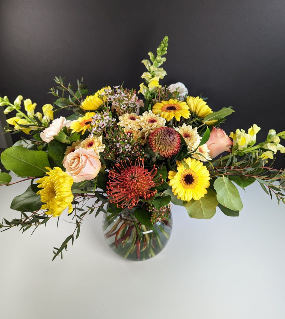 Sunshine Flower Arrangement  - Brighten Your Day with Our Sunshine Bouquet  Illuminate any room with the radiant beauty of our sunshine bouquet. This vibrant arrangement includes:  -Pin cushion protea -Yellow gerbera daisies -Pastel orange roses -Yellow chrysanthemums -Yellow snapdragons -Pastel orange carnations  Perfect for uplifting spirits and spreading joy, our sunshine bouquet is sure to bring warmth and happiness to any occasion.